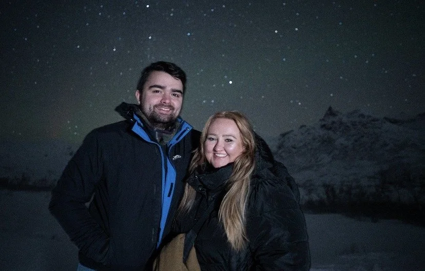 A man and woman outdoors at night, snow-covered mountains in the background, under a starry sky, both wearing winter jackets and smiling.