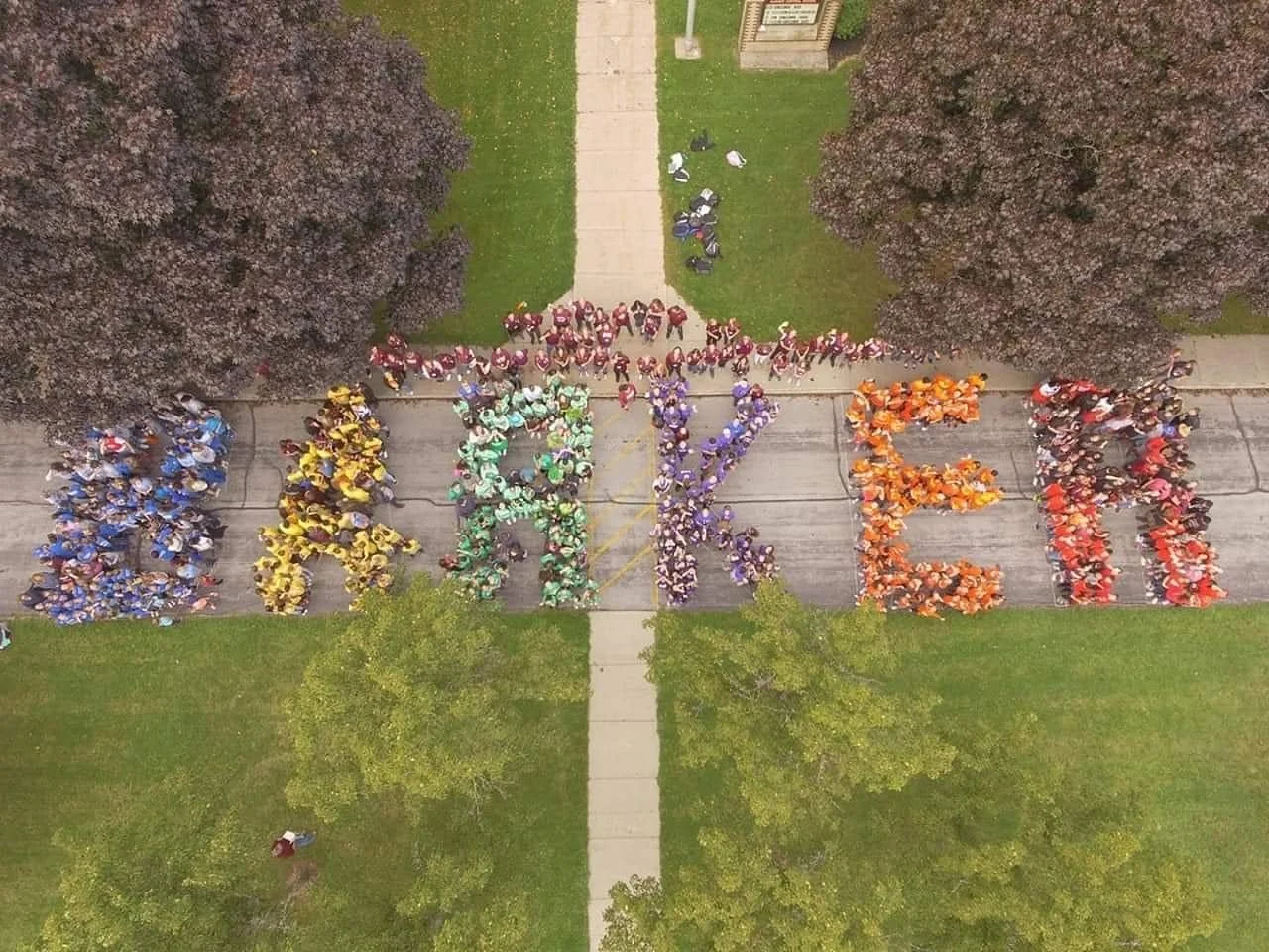 Aerial view of a large group of people forming the word 'AREA' on the ground, with each letter filled with people wearing brightly colored shirts, surrounded by trees and a green lawn.
