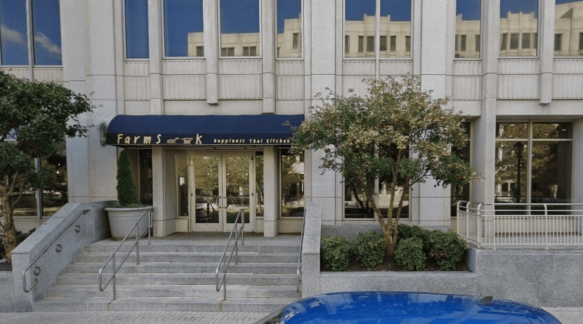 Exterior view of a building with glass windows, a staircase leading to the entrance, and a sign that reads 'Farmers & ck happiness that kitchens.' There are trees and shrubs in front of the building, and a blue car partially visible in the foreground.