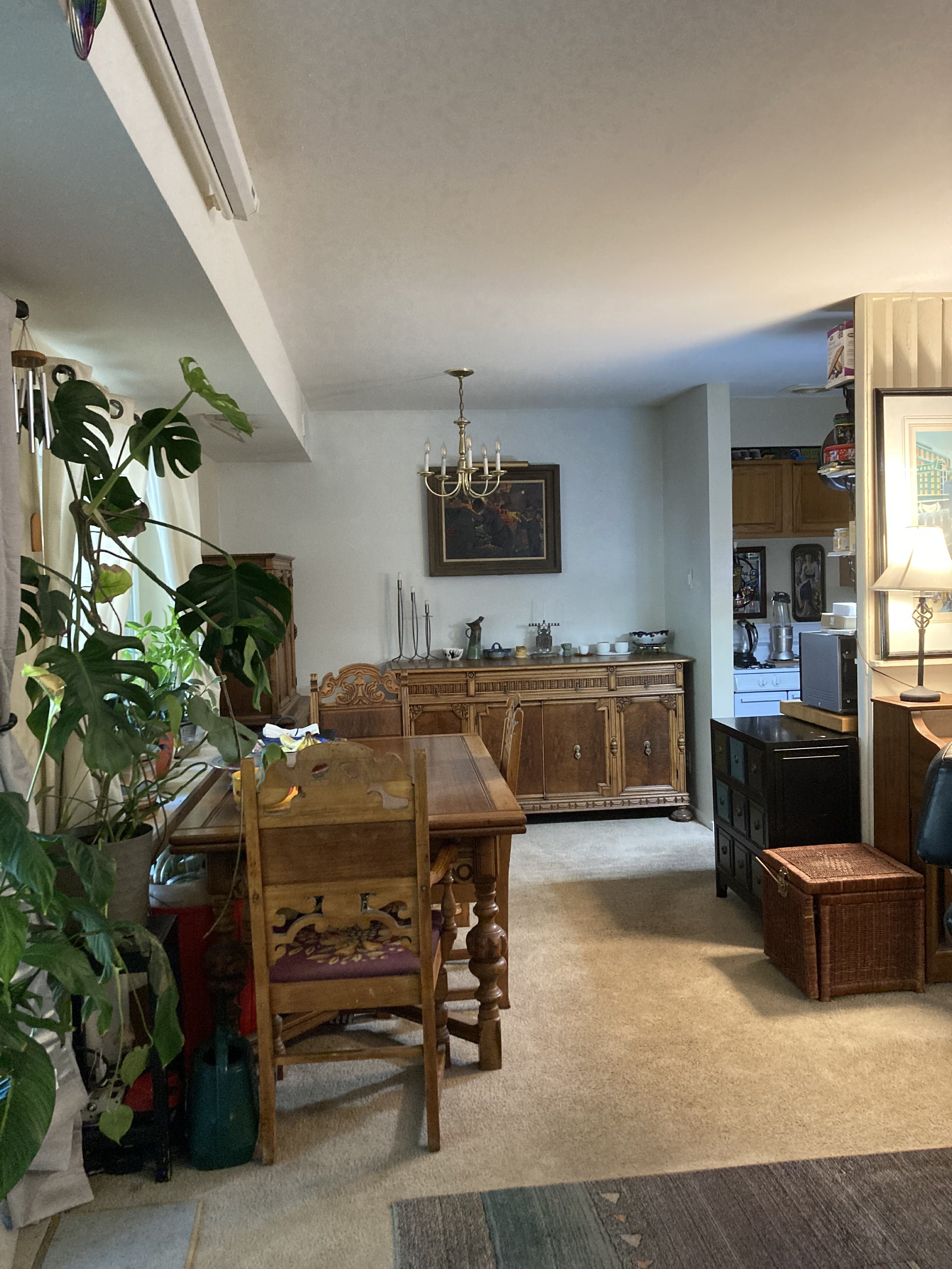 A view of a dining room interior with a wooden dining table and chairs, a sideboard, a chandelier, and a kitchen in the background. There is a large plant on the left, and the room is decorated with artwork and various household items.