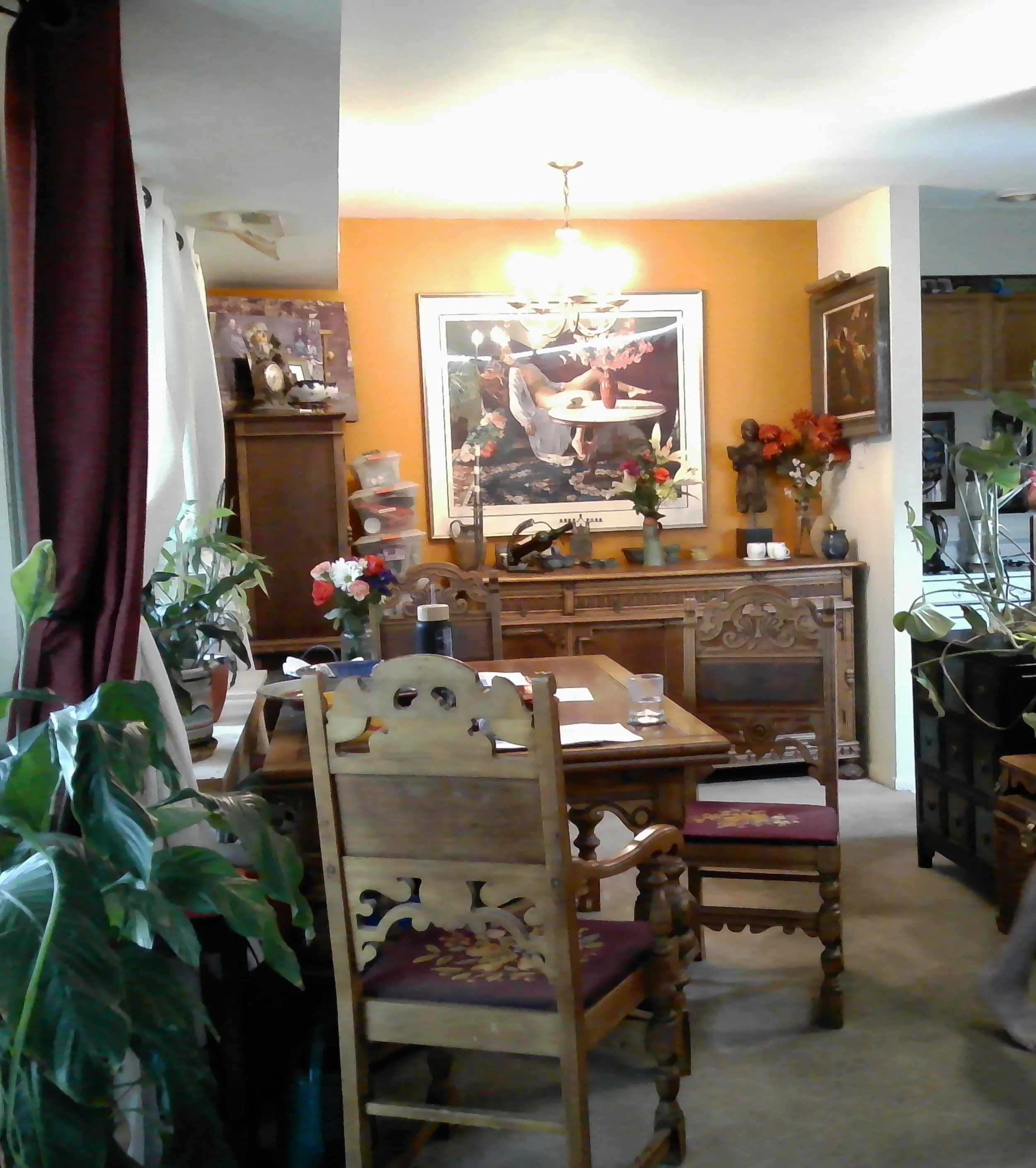 A dining room with a wooden table and chairs, decorated with flowers, framed art on the wall, potted plants near windows with curtains, and a sideboard with vases and ornaments.