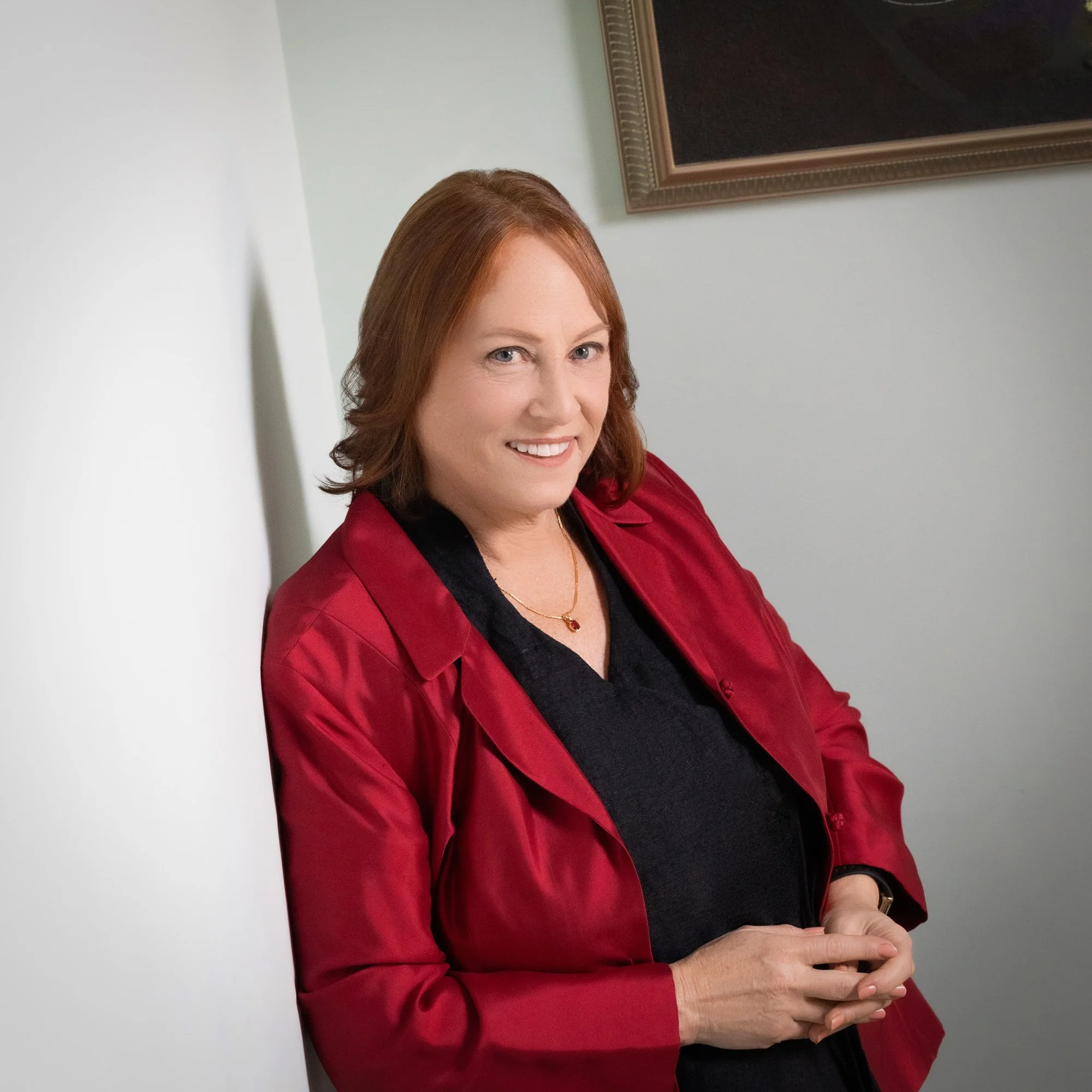 A woman with reddish-brown hair, wearing a red blazer and black top, smiling and leaning against a white wall in an indoor setting with a framed picture on the wall.