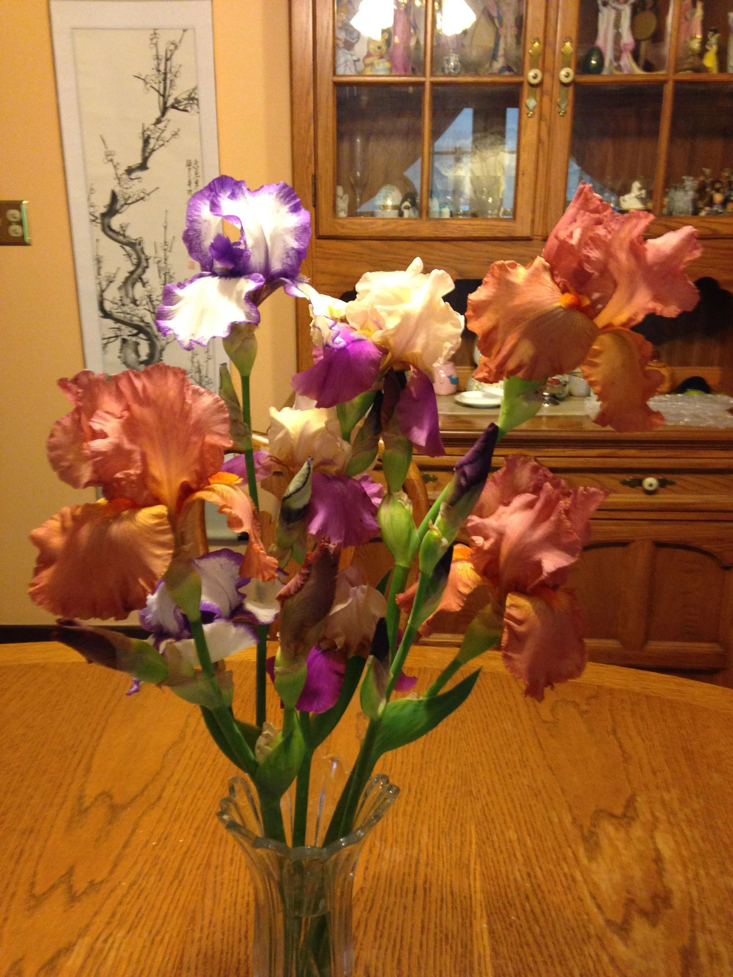 A bouquet of colorful irises in a glass vase on a wooden table in a dining room. In the background, a wooden cabinet with glass doors displays decorative items, and a wall hanging with a black ink drawing of a tree is visible.