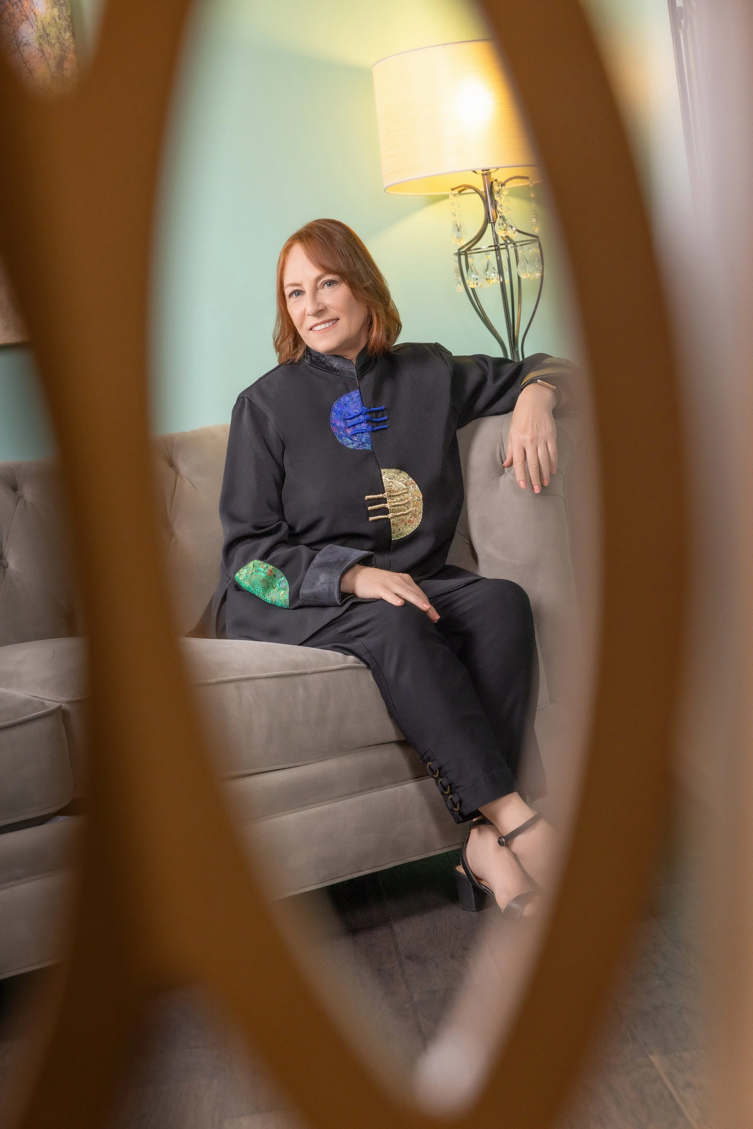 A woman with shoulder-length red hair, wearing a black jacket with colorful circular patterns, sitting on a beige sofa, smiling, with a table lamp behind her, viewed through a decorative wooden cutout.