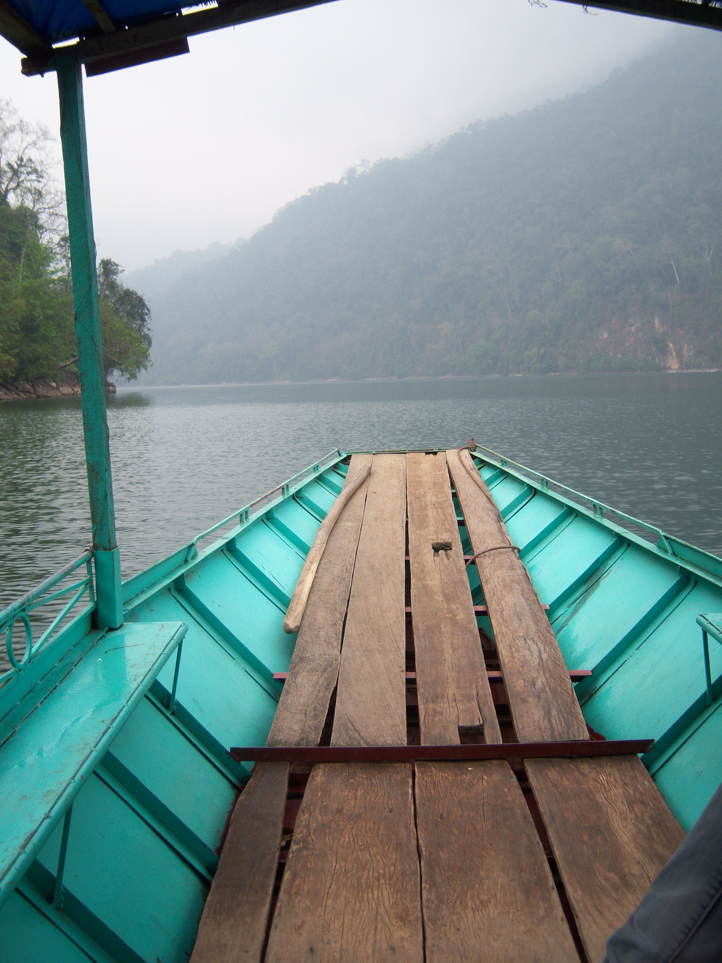 View from a boat on a misty lake with a wooden dock leading into the water, surrounded by lush green hills.