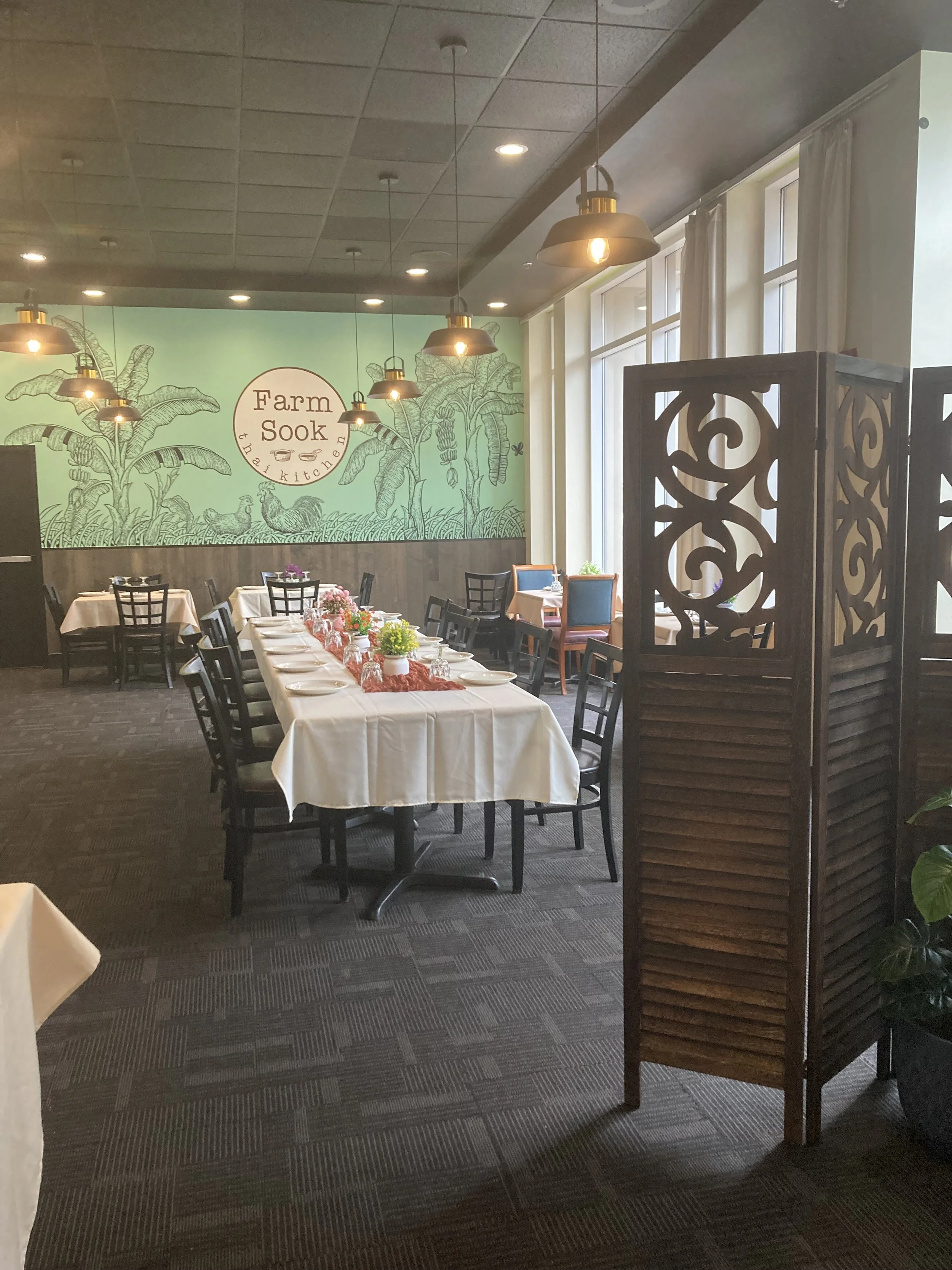 Inside a restaurant with a long table set for dining, decorated with potted plants and pink table runner. There are several black chairs around the table and a wooden decorative room divider on the right. A large window with white curtains allows natural light, and a wall mural displays 'Farm Sook Thai Kitchen' with illustrations of plants and chickens.