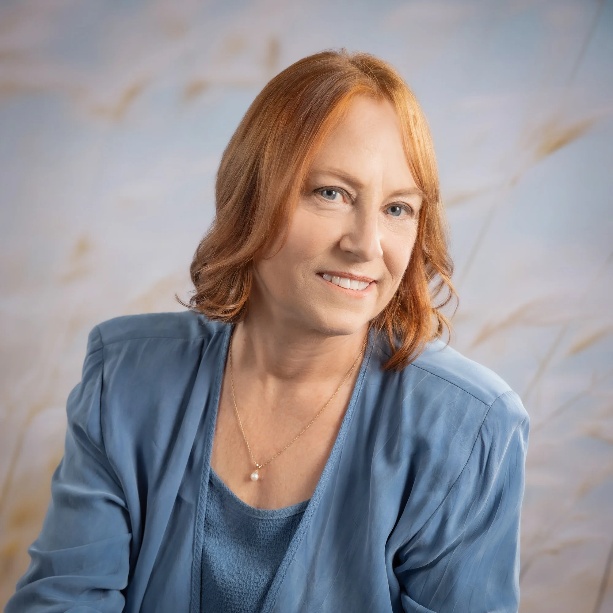 A woman with red hair smiling, wearing a light blue blazer and a pearl necklace, against a soft, blurred background with beige and blue tones.