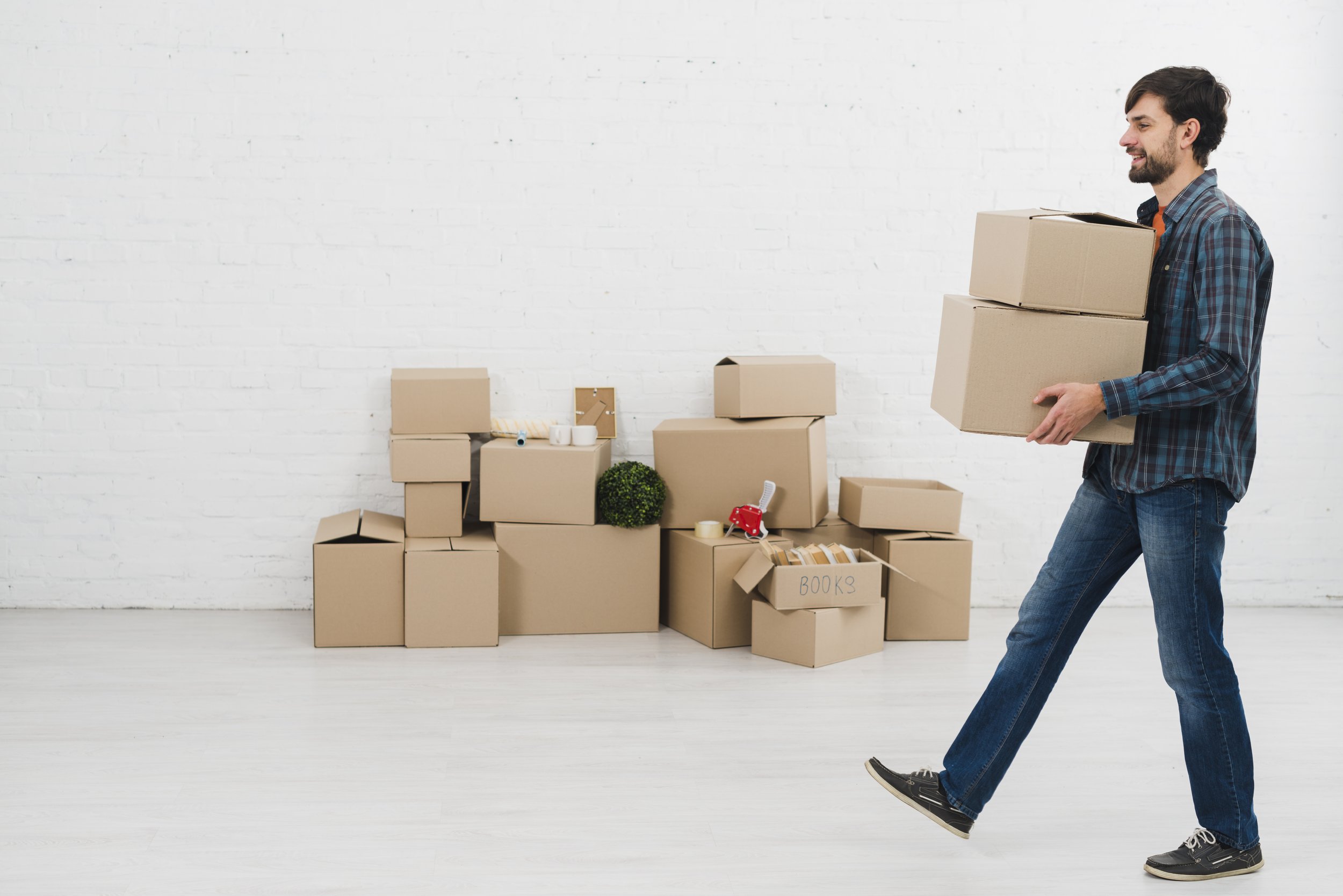 A man carrying a cardboard box in a room with a white brick wall, with packed boxes, tape, and small decorative items on the background floor.