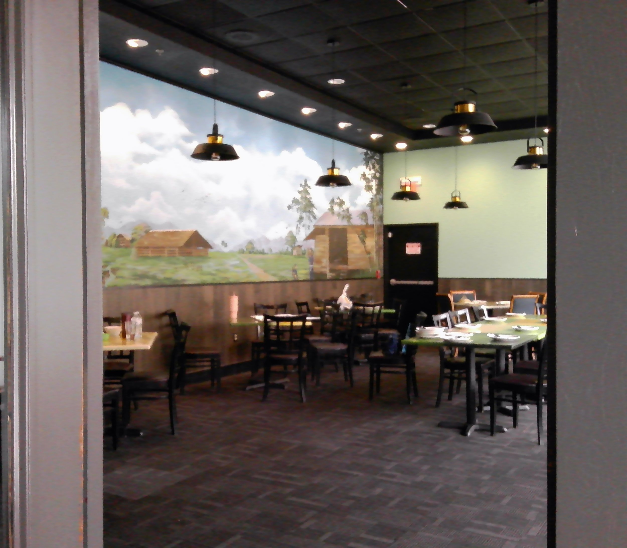 Empty restaurant with wooden tables and black chairs, wall mural of rural farm scene, black ceiling with hanging lamps, and a green accent wall.