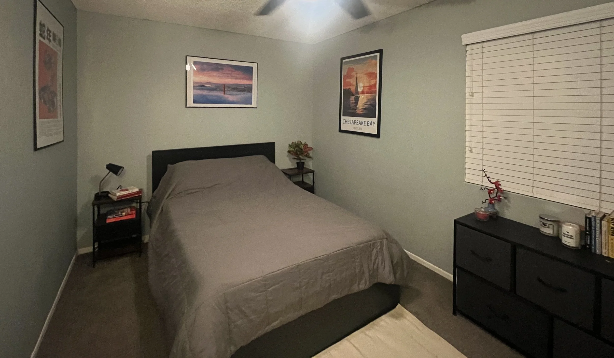 A bedroom with a bed having a gray comforter, framed wall art above it, a nightstand with books and a lamp, and a black dresser with decorative items near a window with blinds.