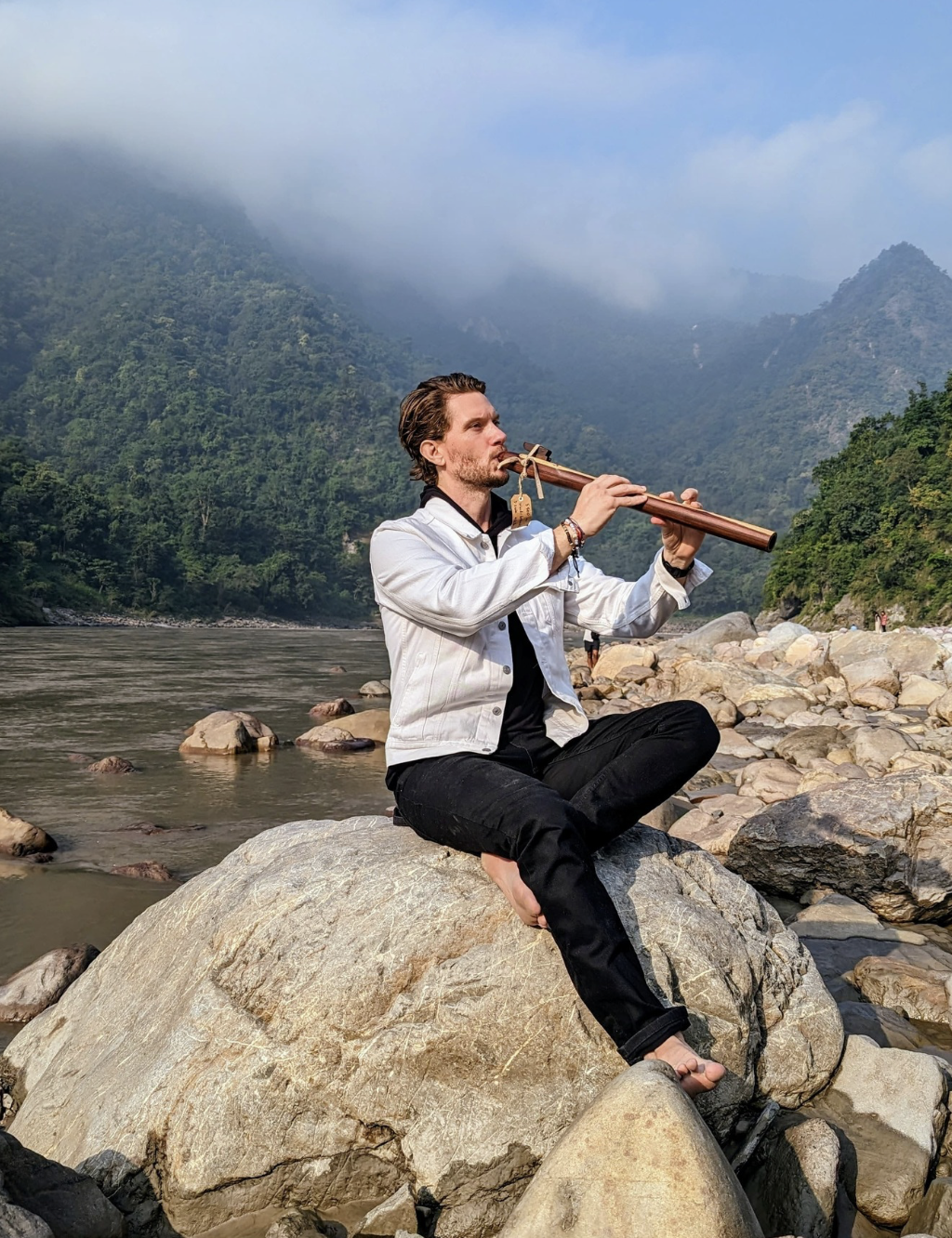 A man sitting cross-legged on large rocks by a river in a mountain landscape, playing a flute, with fog-covered mountains in the background.