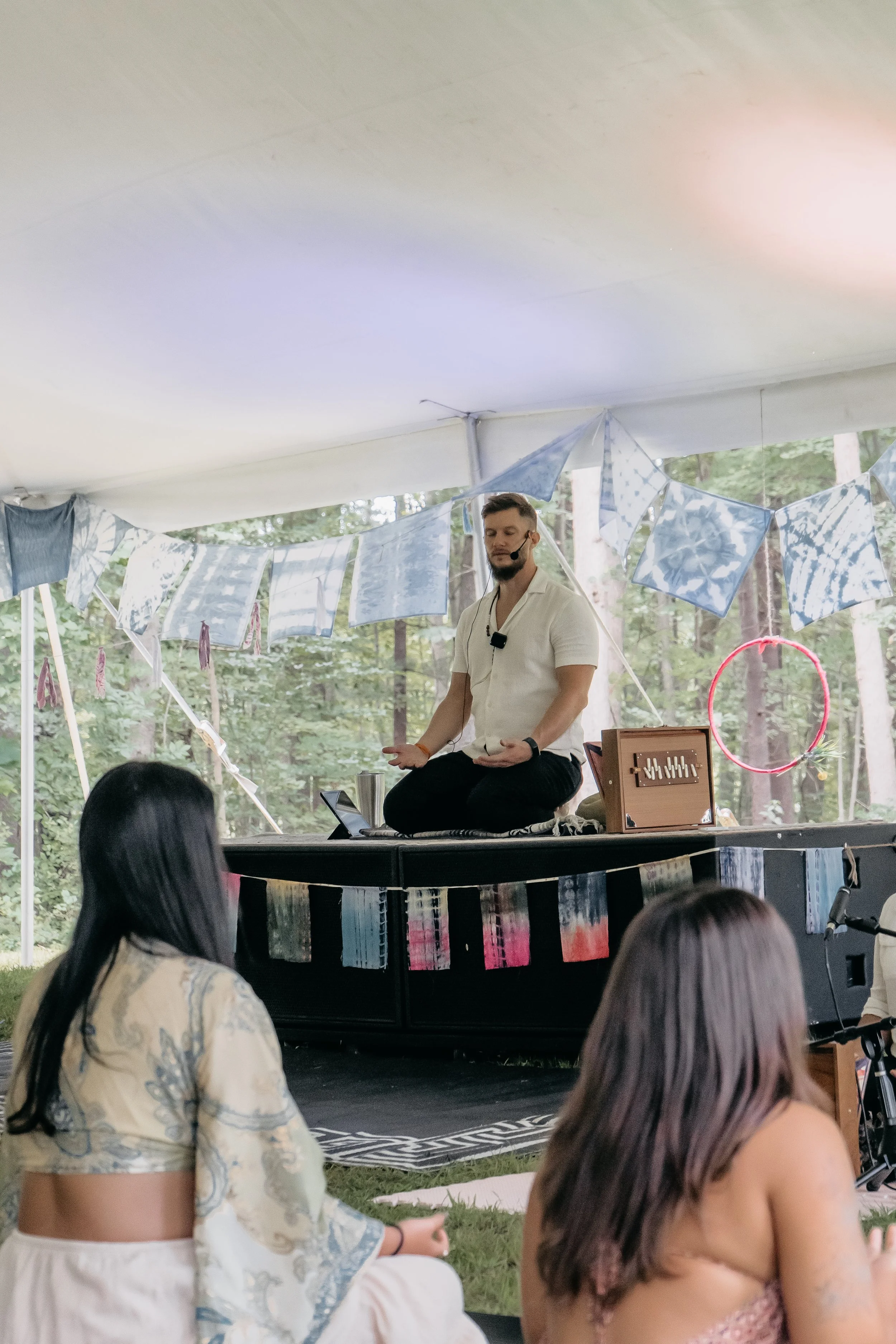 A man sitting on a platform or stage, engaged in a meditative or spiritual activity, with a microphone headset. Two women sitting on the grass are attentively watching him, in an outdoor setting with trees and decorative fabric banners hanging overhead.