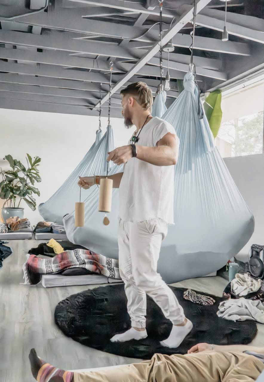 A man in white pajamas practicing meditation with hanging chimes inside a room with a hammock in the background and various clothing and objects scattered on the floor.