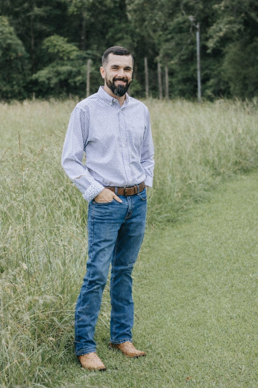 A man with a beard smiling outdoors wearing a checkered shirt, jeans, and tan shoes, standing in a grassy field with trees in the background.