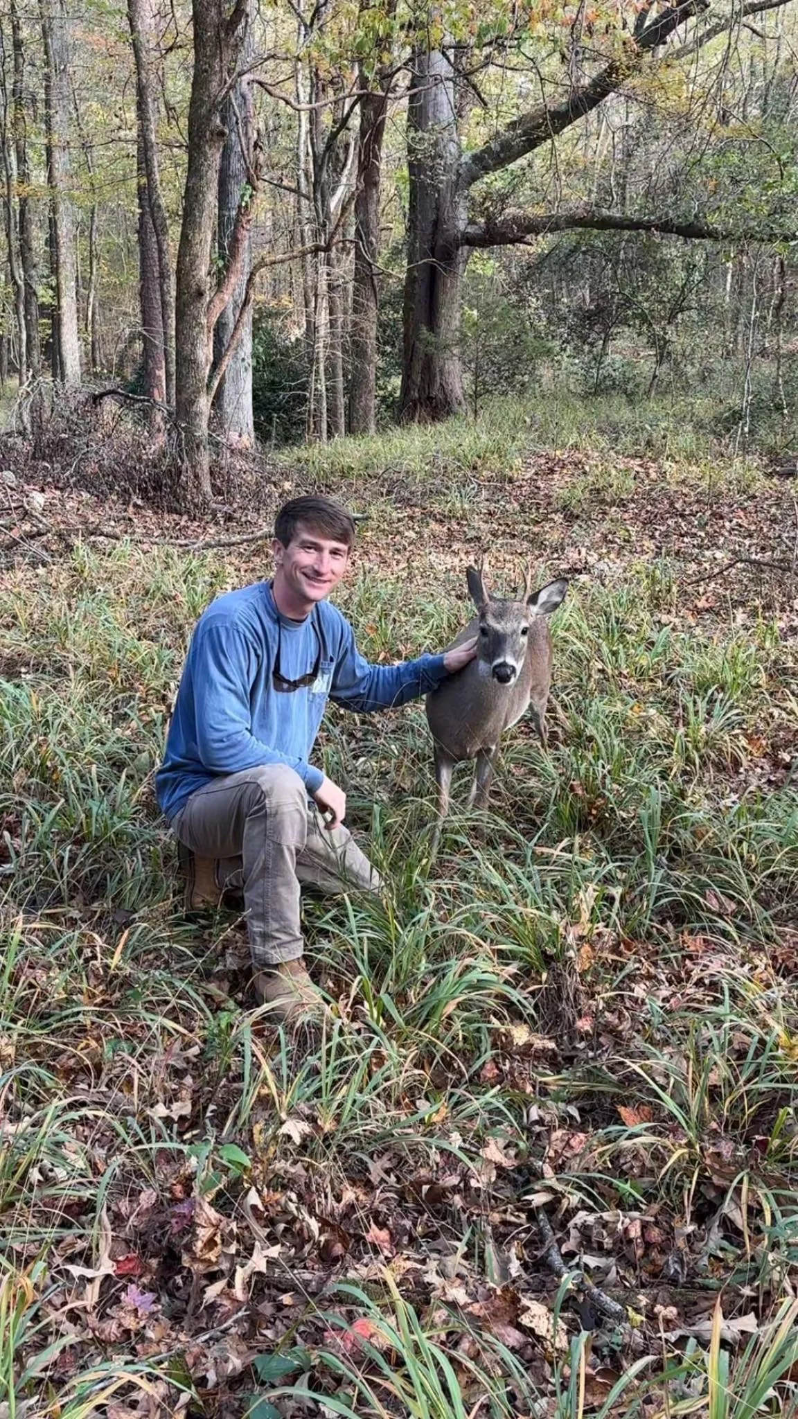A man wearing a blue long-sleeve shirt and beige pants kneeling in a forest clearing with a young deer.