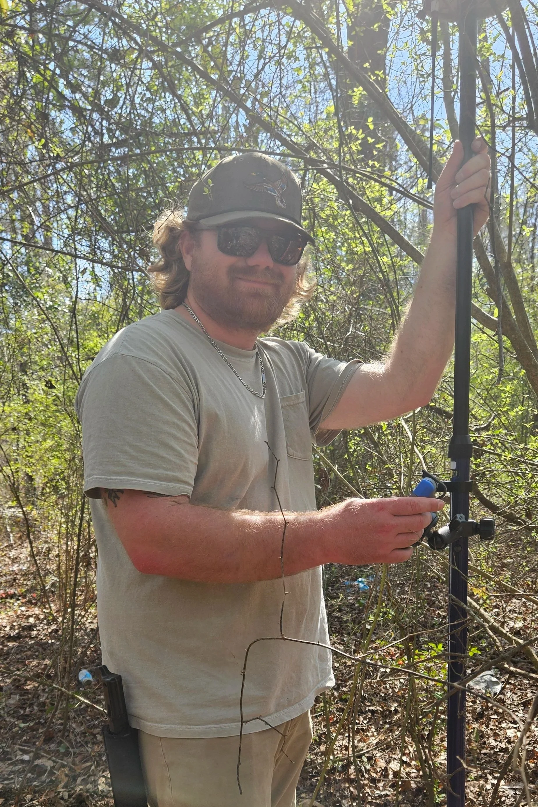 A man in sunglasses, a beige t-shirt, and a baseball cap, standing in a wooded area, holding a metal detector.