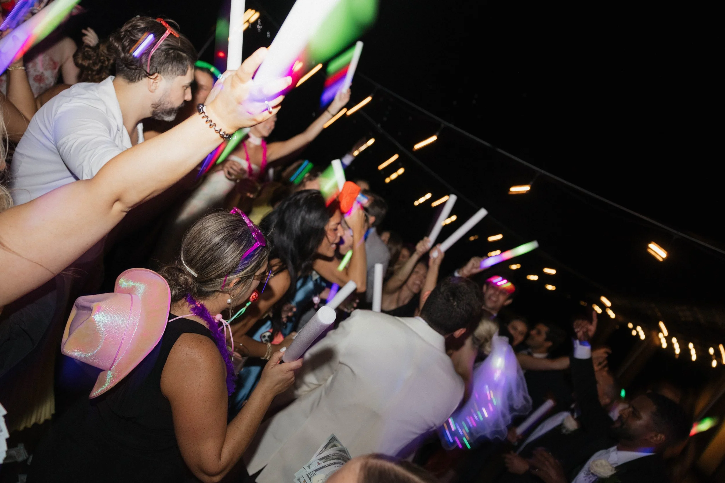 Decorated wedding cake with a wedding topper of a couple dancing, surrounded by a variety of flowers, on a table set with wine glasses, plates, and utensils, with a wallpaper featuring a pastoral scene in the background.