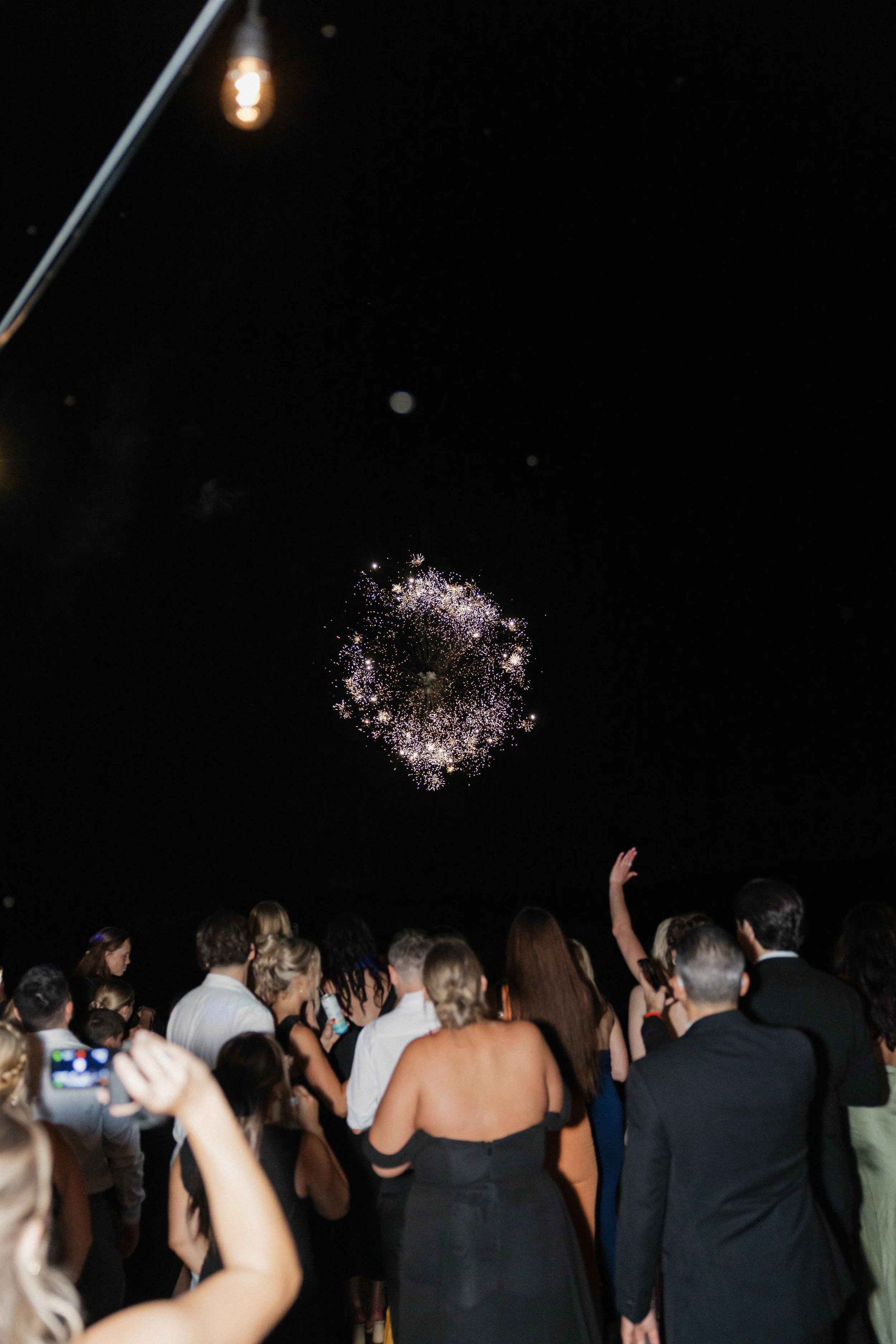 A flower arch decorated with colorful flowers stands in front of a red wooden fence on a grassy lawn, with small white chairs arranged on either side for a wedding or event.