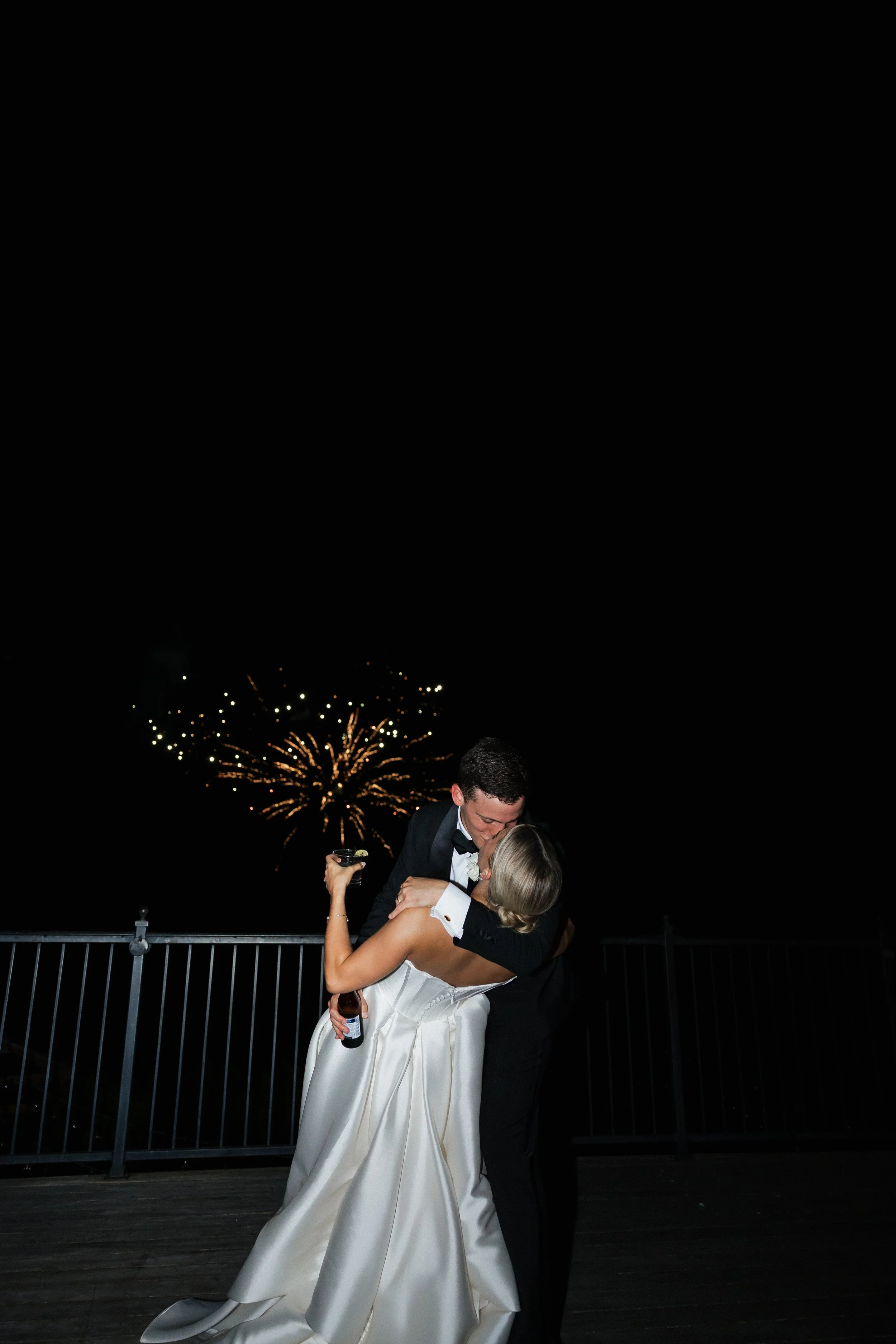 A newlywed couple stands outdoors, smiling and holding hands, with the groom leaning in to kiss the bride's forehead. The bride wears a lace wedding dress and a floral crown, while the groom is dressed in a dark suit and bow tie. Background includes greenery and blurred plants.