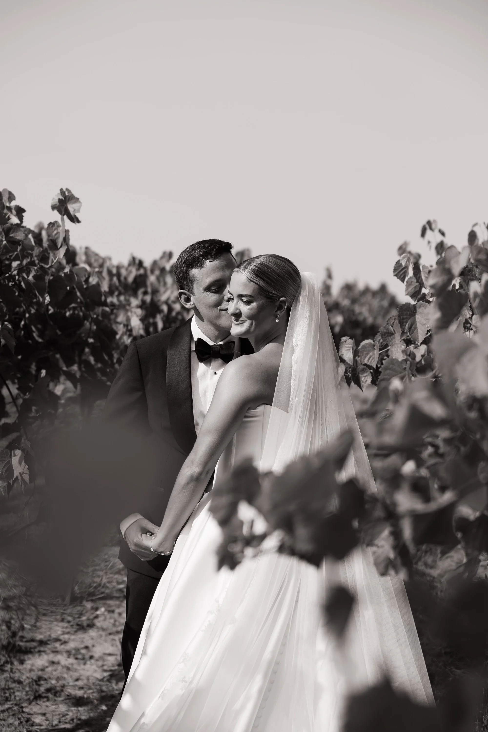 Elegant outdoor event setup with a long, white-clothed table decorated with greenery and white floral arrangements. Hanging lanterns and string lights with paper lanterns are overhead. The scene is surrounded by lush green trees.