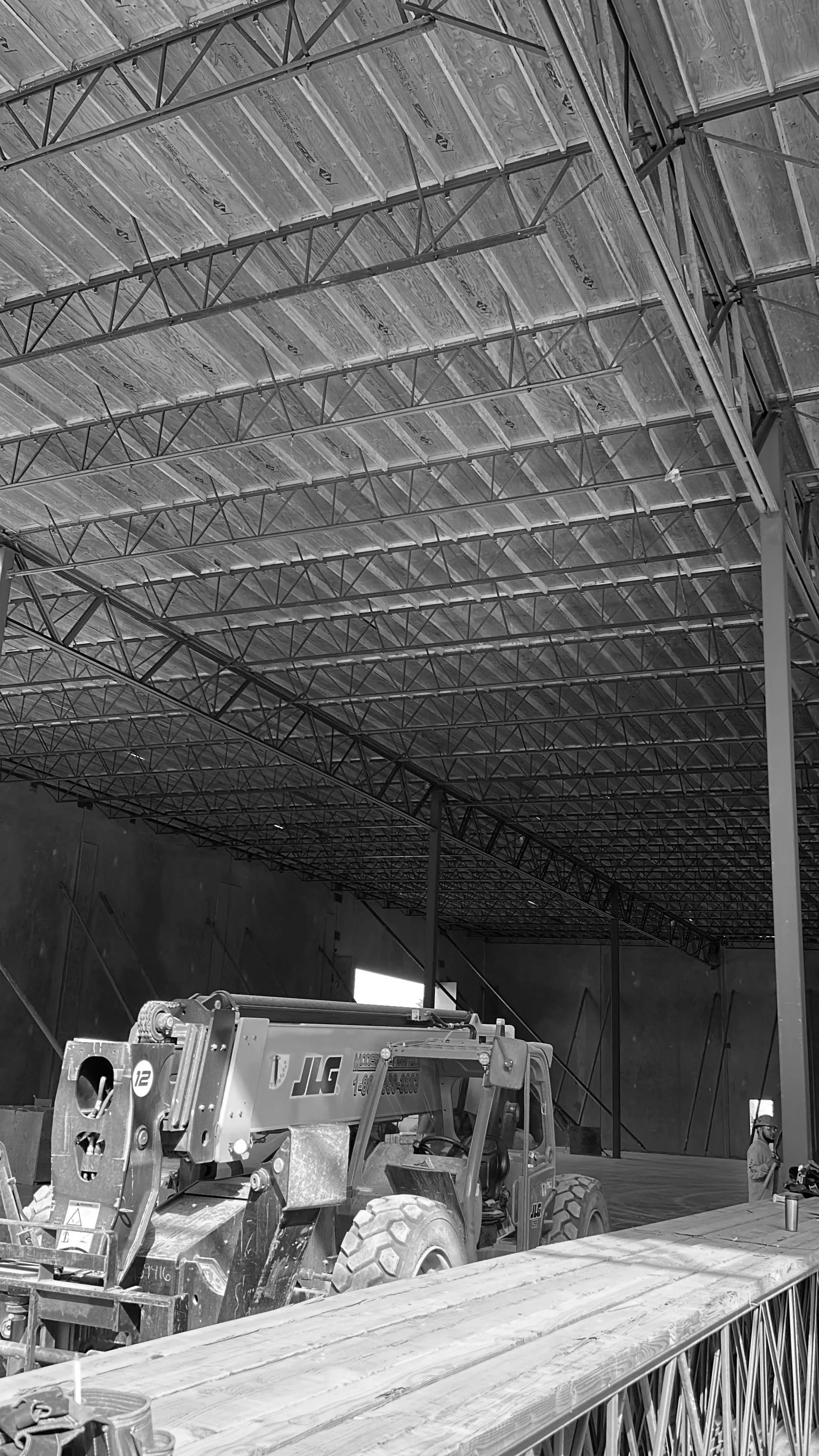 Construction site with steel framework and a JLG crane, worker in safety helmet and vest, and wooden beams in the foreground.