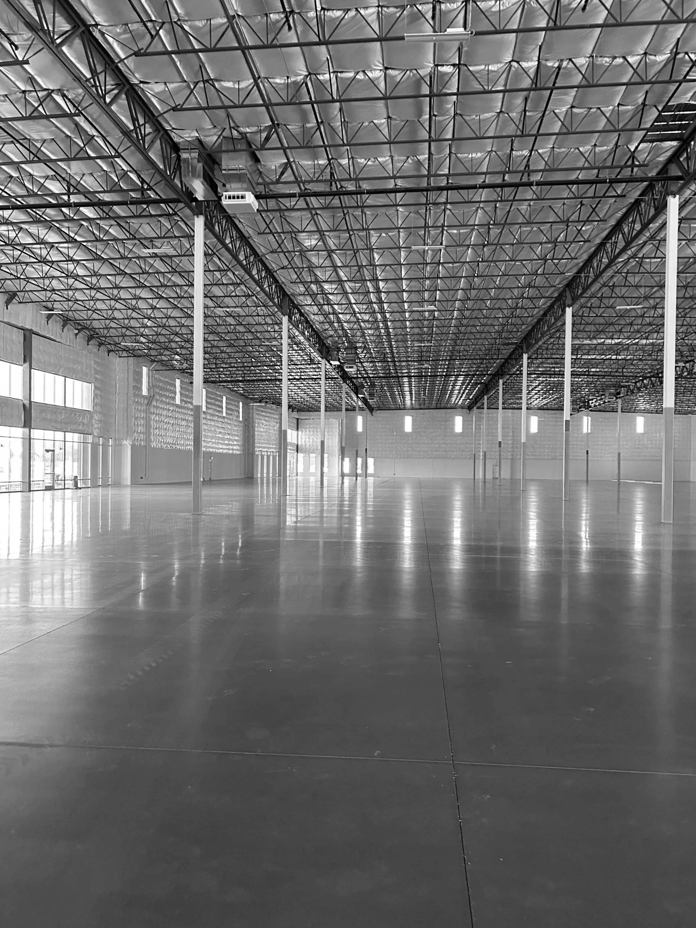 Empty indoor gymnasium with polished floor, large windows on the side, and a high ceiling with metal trusses and insulation.