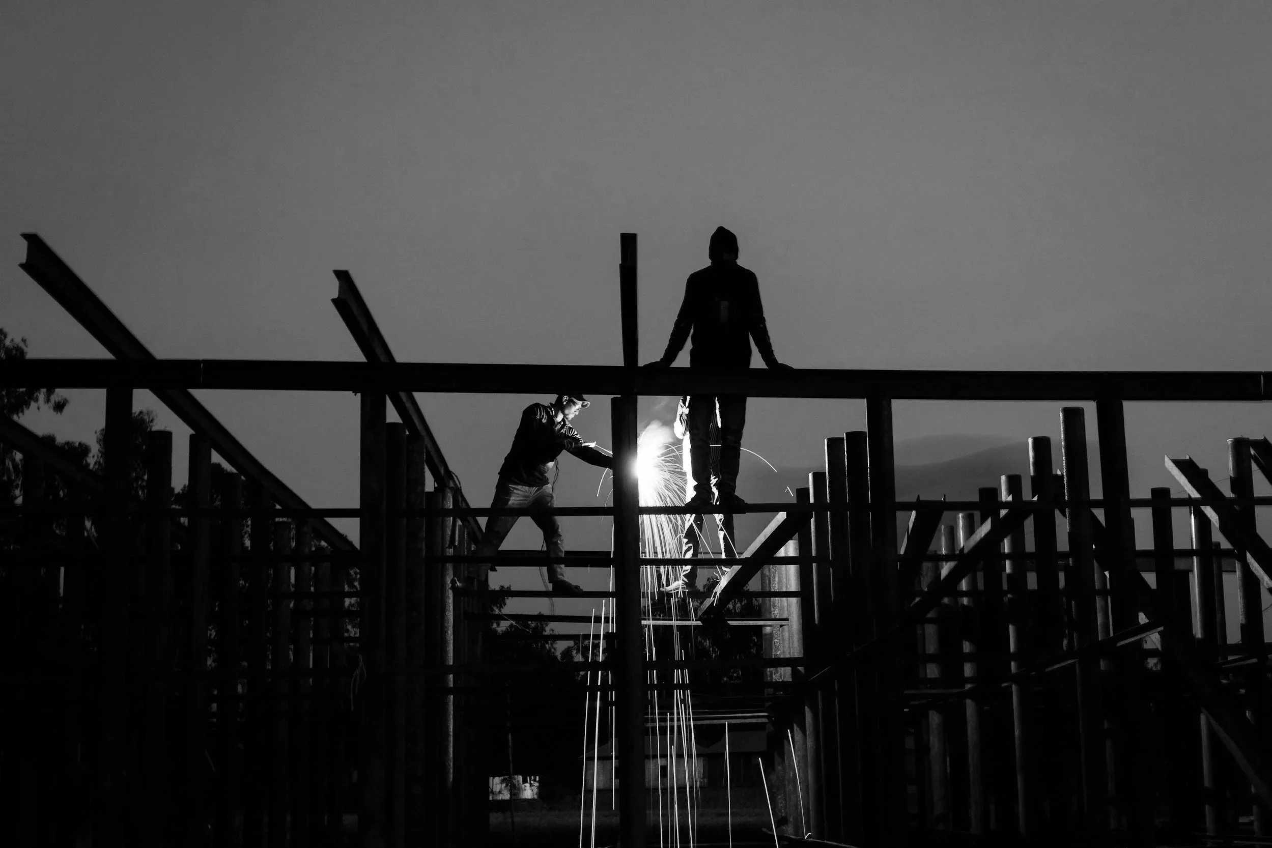 Silhouettes of two construction workers welding on a wooden framework during dusk or nighttime, with sparks flying.