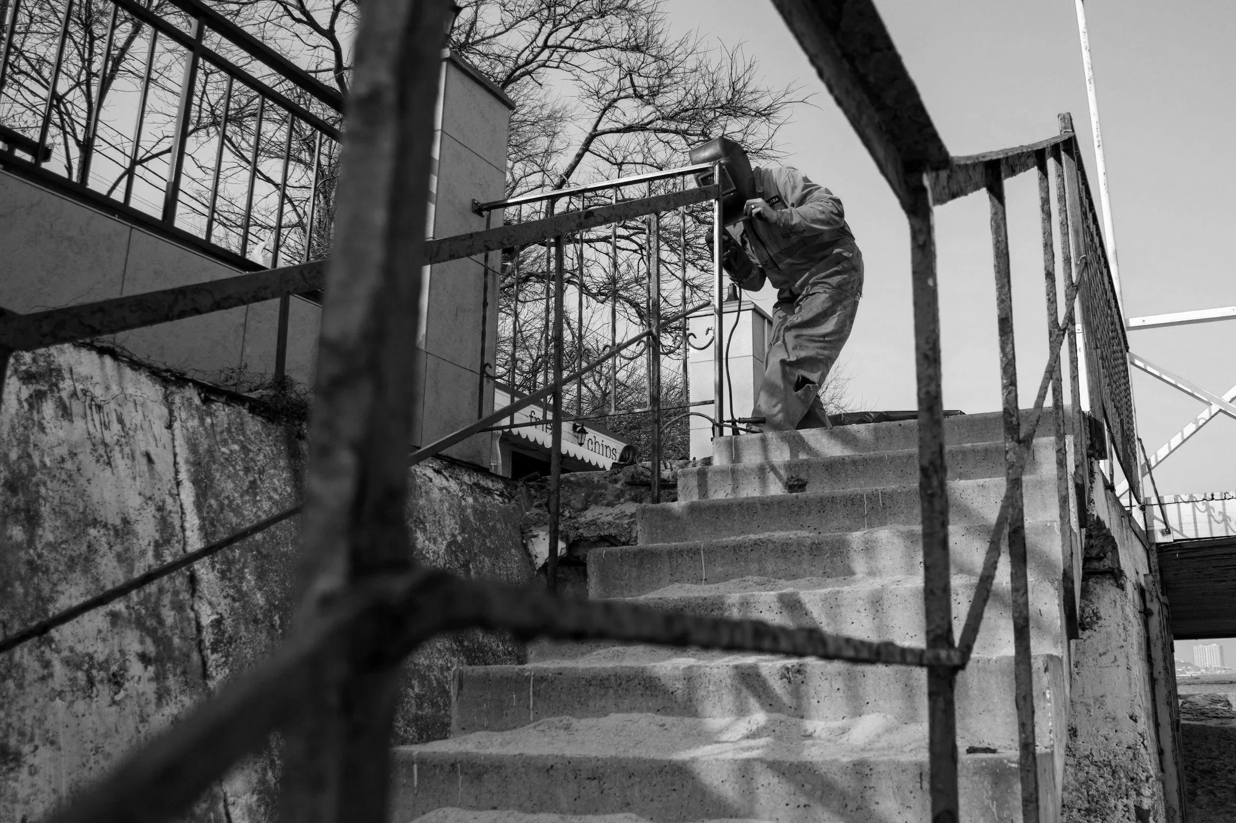 A worker with a welding helmet and work clothes welding on a railings outdoors on stairs, with trees and sky in the background, in black and white.