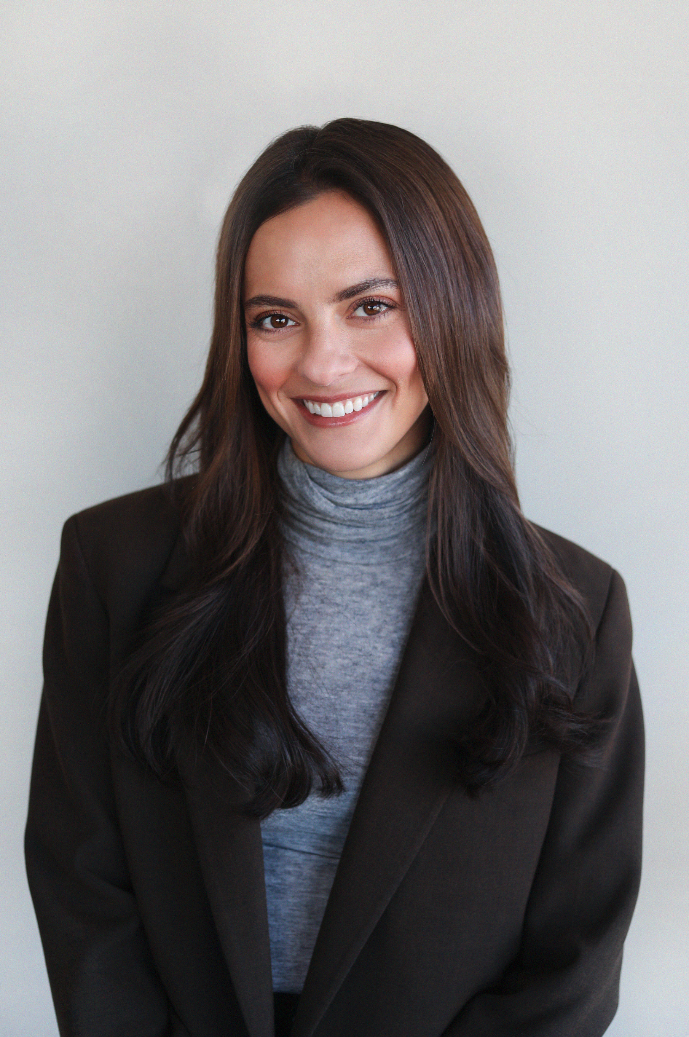 A woman with long dark brown hair, smiling, wearing a gray turtleneck and a dark blazer, against a plain light gray background.
