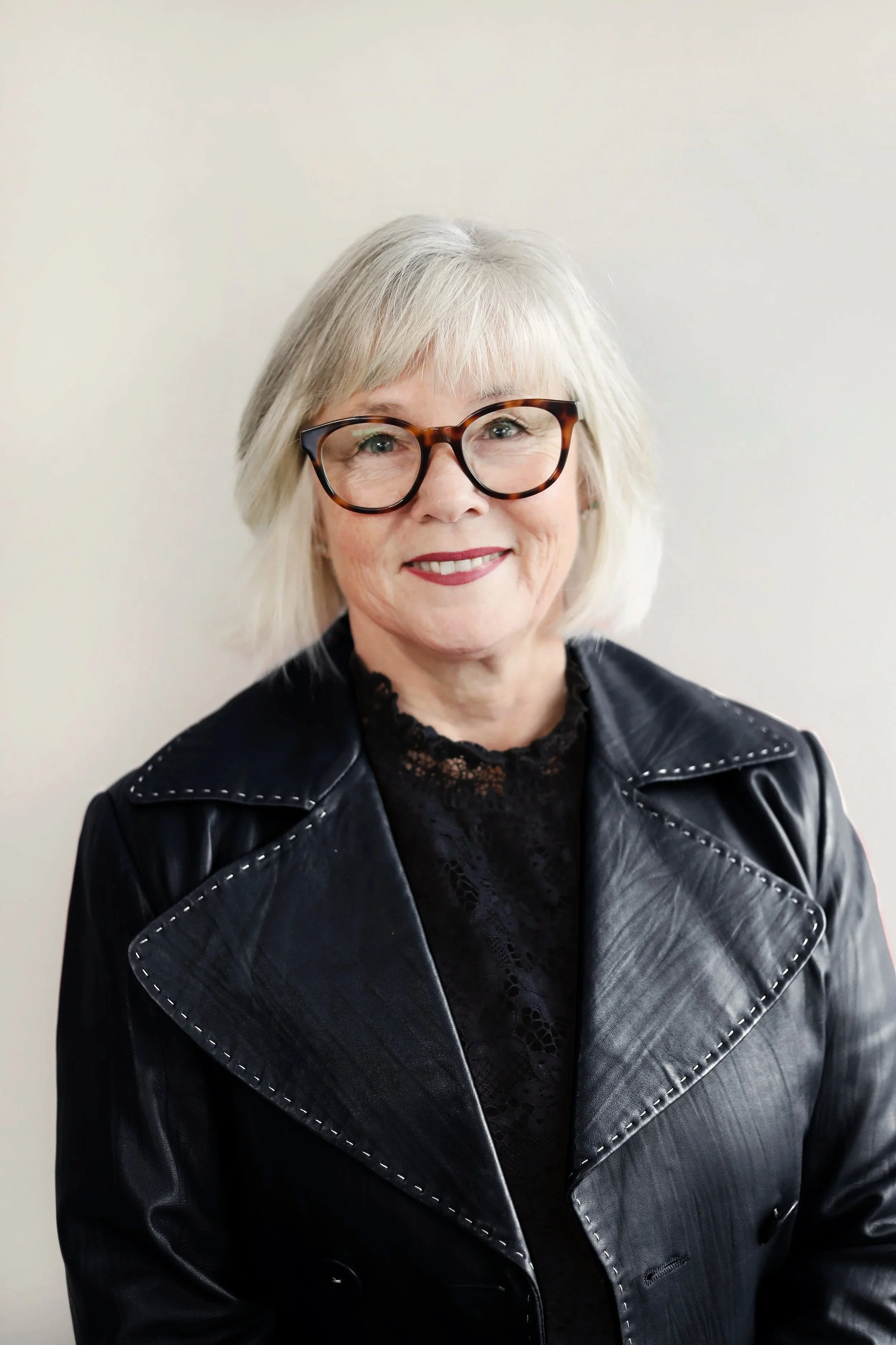A woman with short gray hair and tortoiseshell glasses smiling, wearing a black leather jacket over a lace black top, standing against a plain light-colored background.