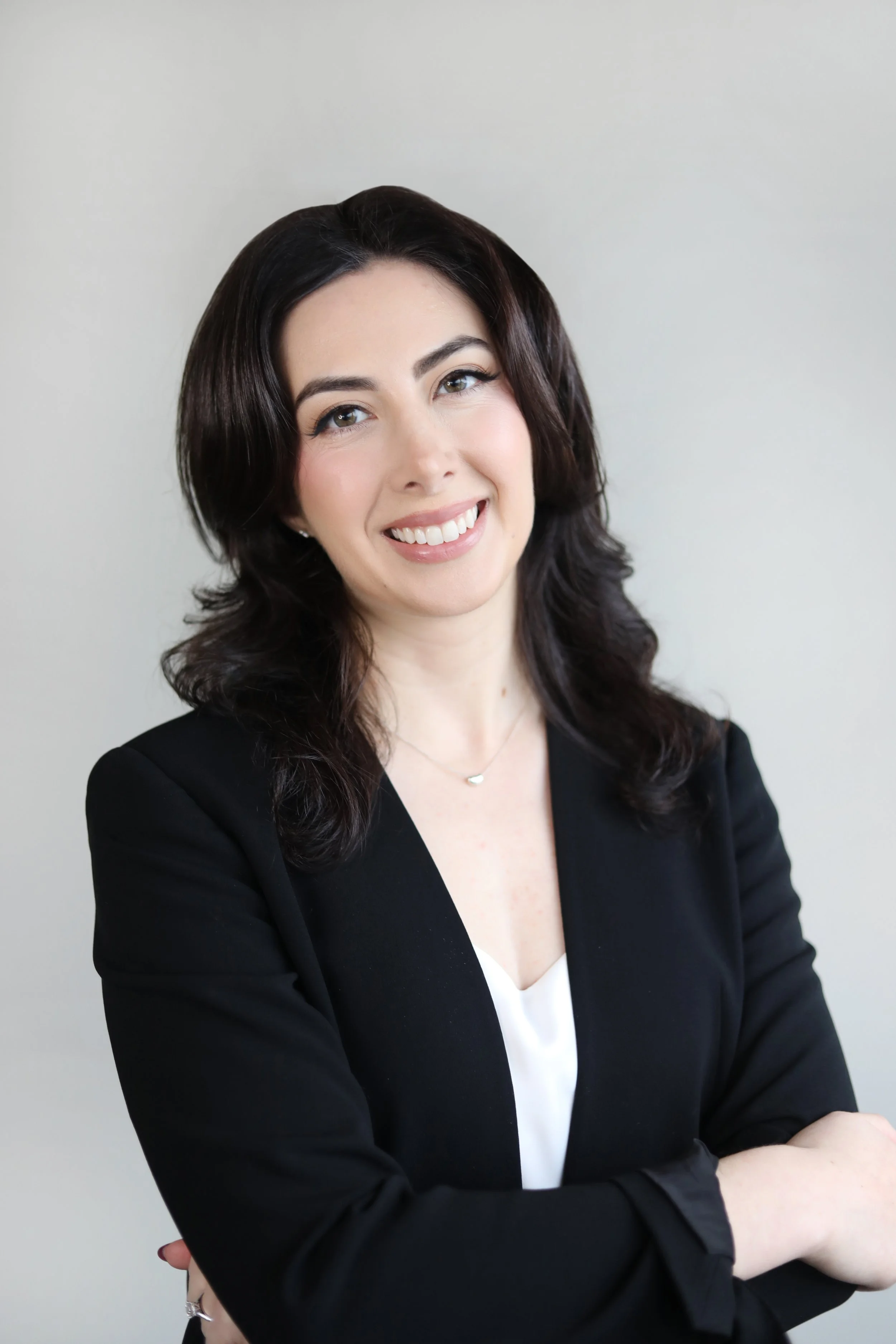 A woman with dark, wavy hair and light skin, smiling, wearing a black blazer over a white top, against a plain light-colored background.