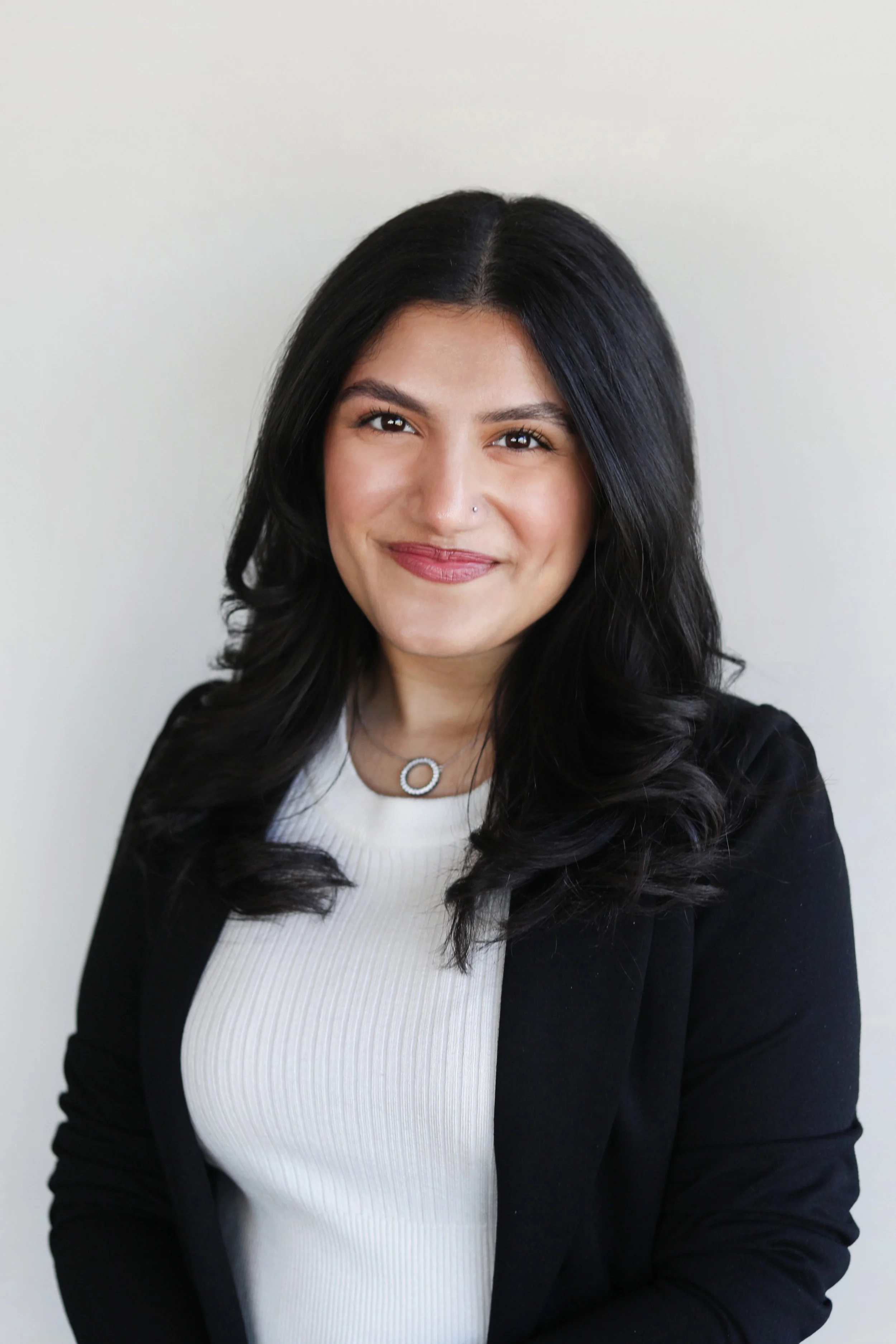 Headshot of a woman with long dark wavy hair, wearing a white top and black blazer along with a circular pendant necklace, smiling in front of a plain light background.