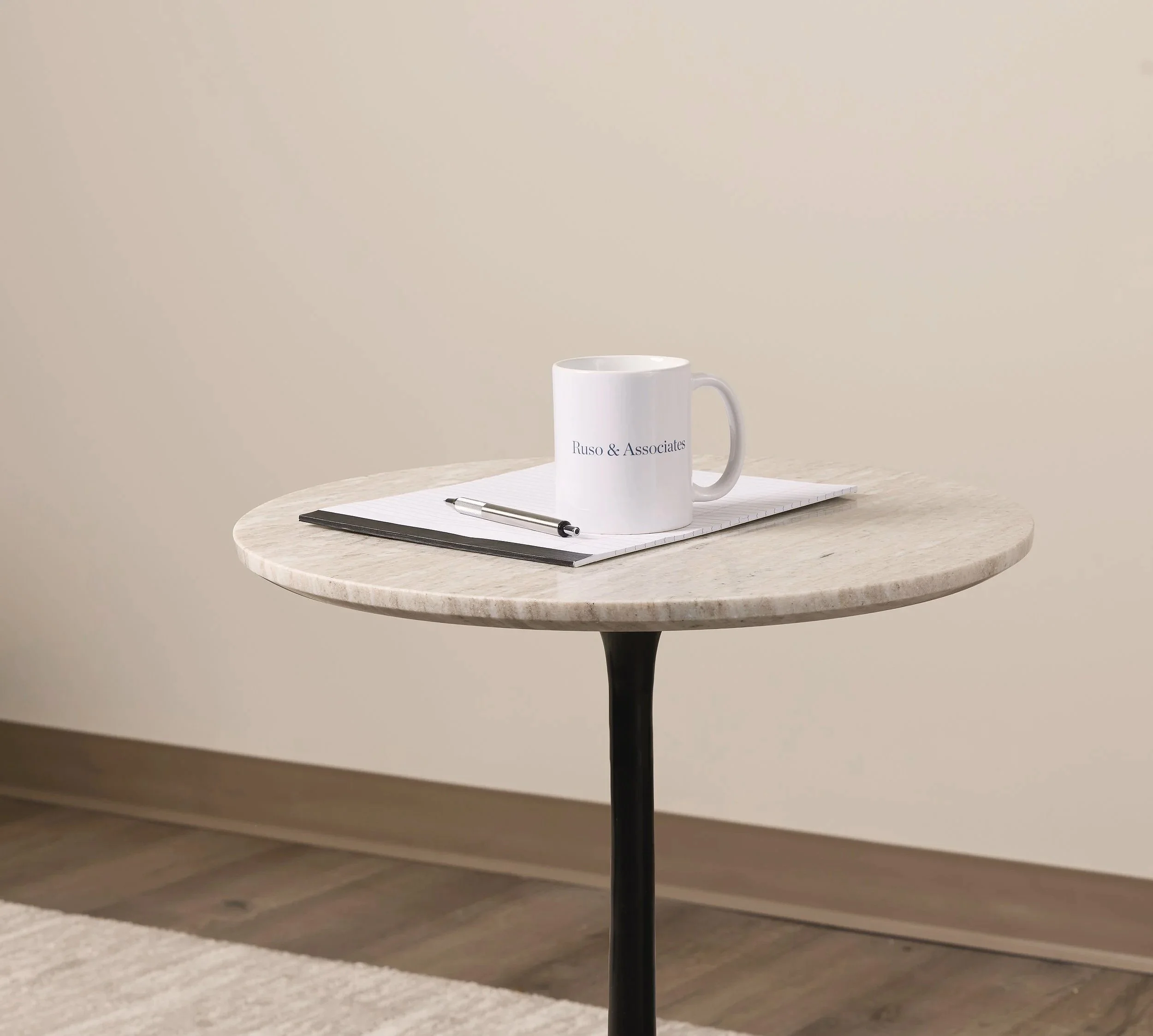 A white ceramic mug with the text 'Ruso & Associates' printed on it, placed on a white paper notepad on a round marble table with a dark base, set against a plain beige wall.