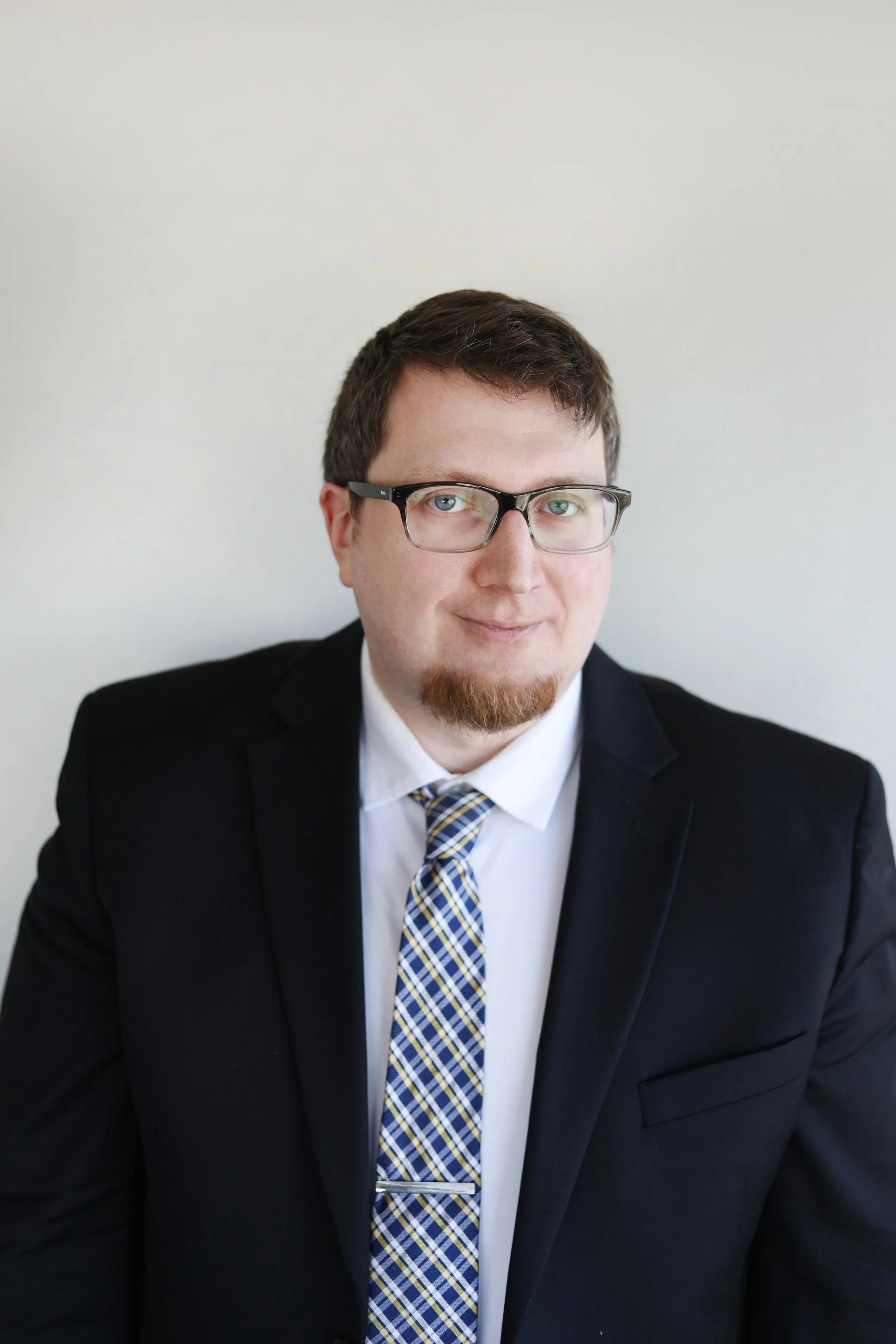 Young man with brown hair, wearing glasses, a dark suit, white shirt, and a checkered tie, smiling against a plain light-colored wall background.