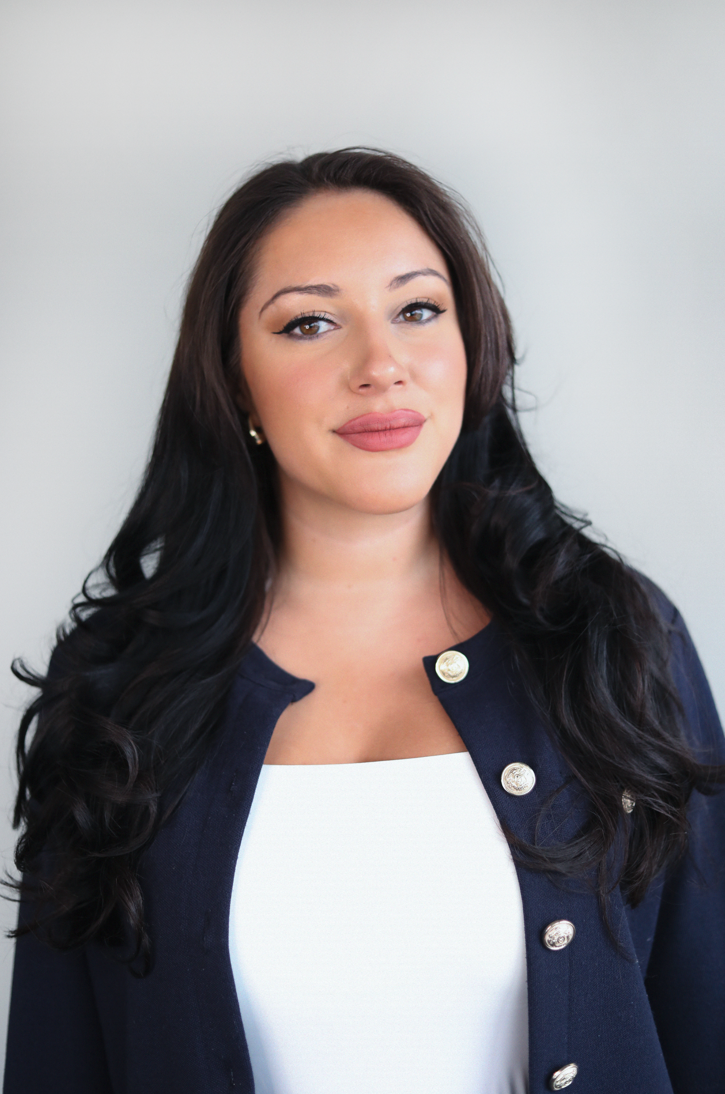A woman with long dark hair, light makeup, and wearing a white top and a navy blazer with silver buttons, standing against a plain light background.
