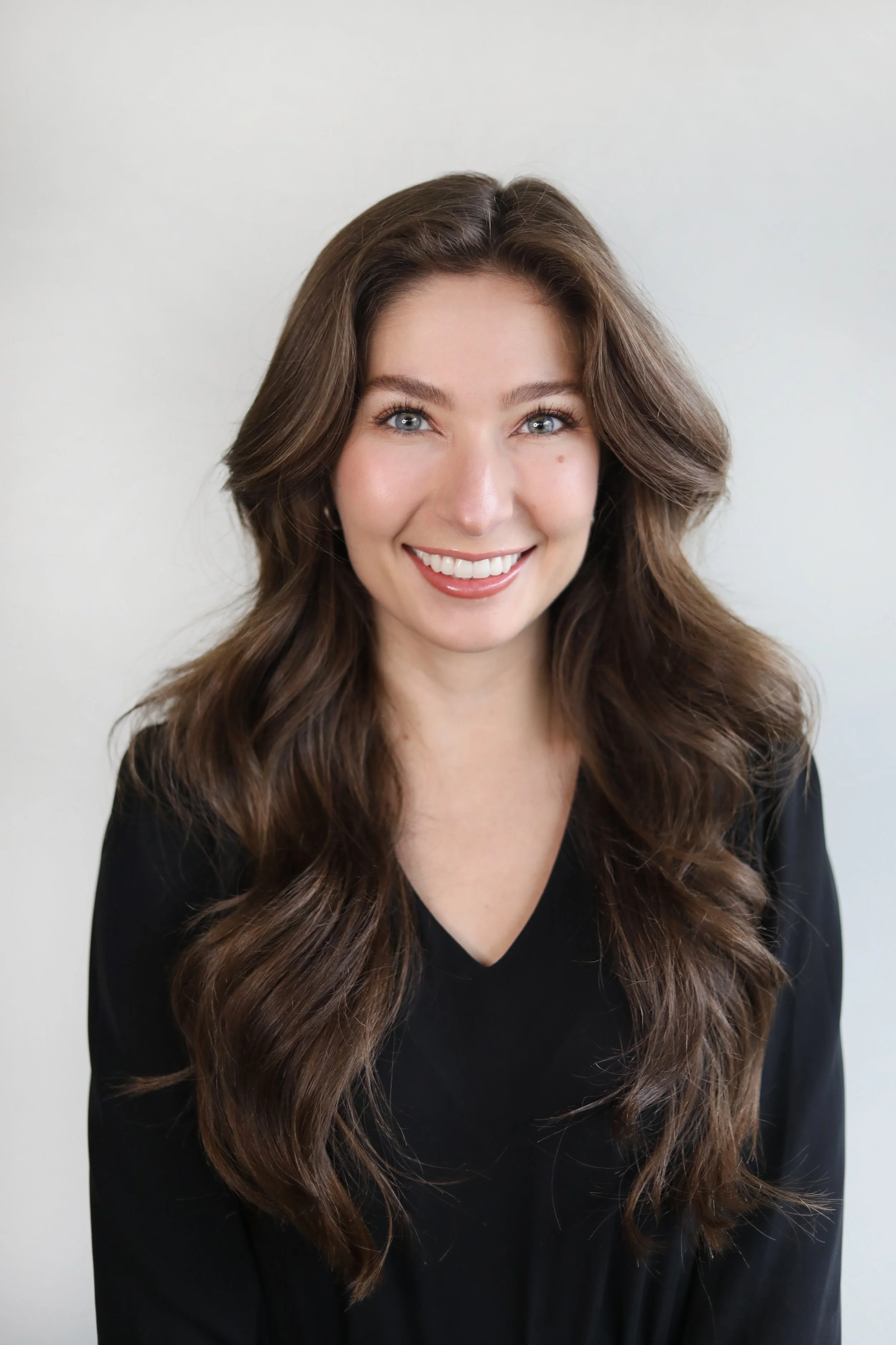 Portrait of a woman with long, wavy brown hair, blue eyes, and a bright smile, wearing a black top, standing against a plain white background.