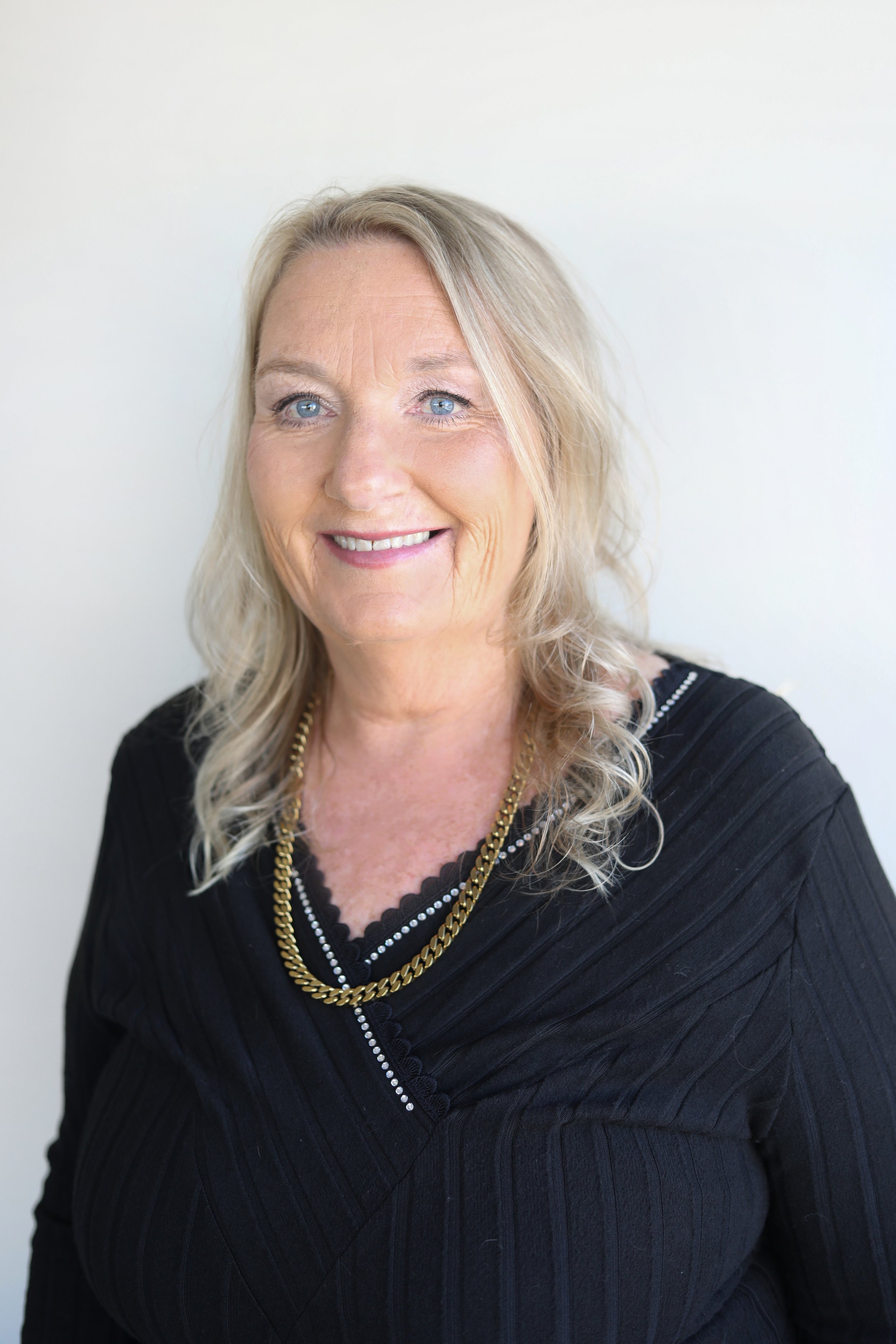 A woman with blonde hair and blue eyes smiling in front of a plain white wall, wearing a black top and layered gold necklaces.