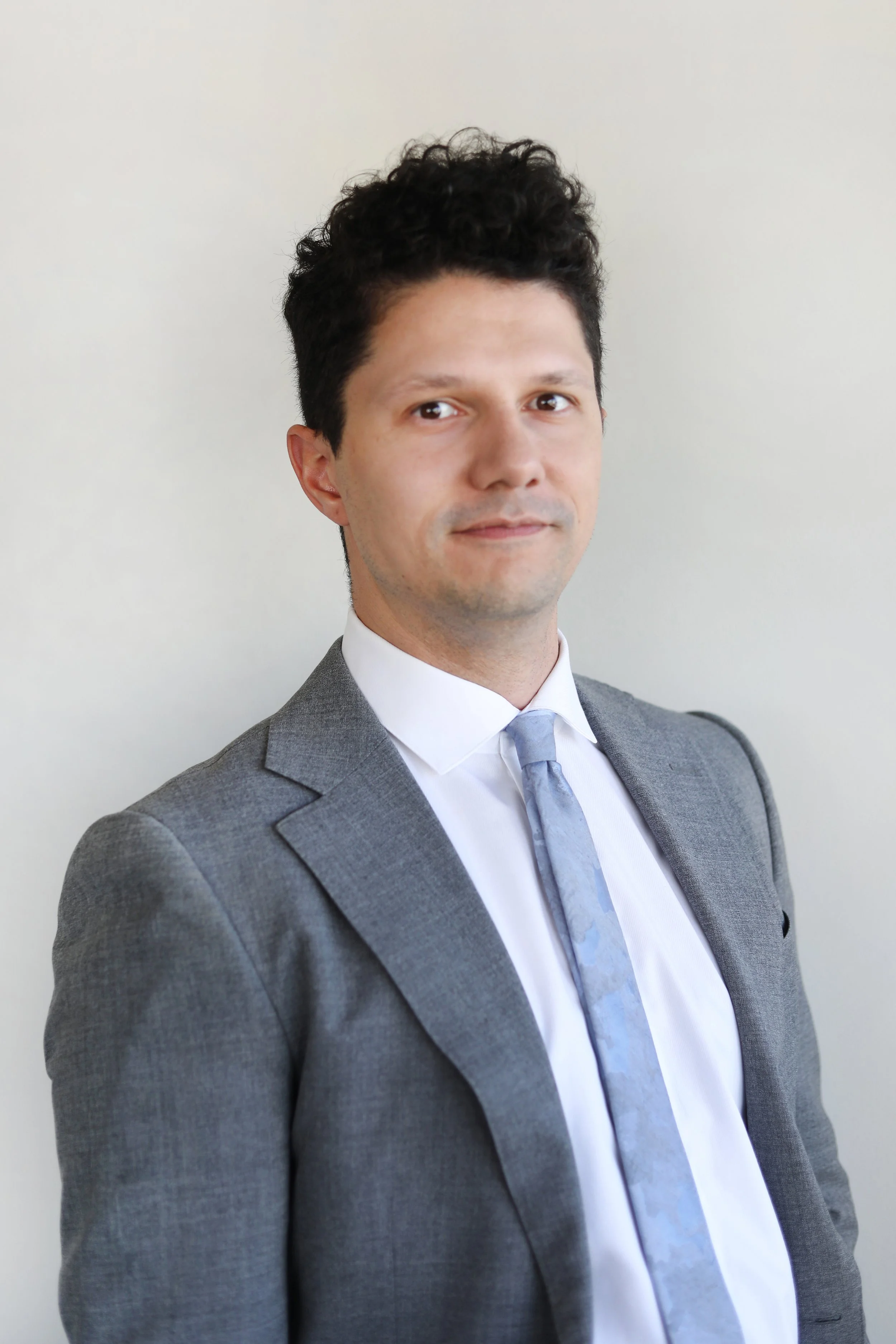 A man in a gray suit, white shirt, and light gray tie standing against a plain light-colored wall.