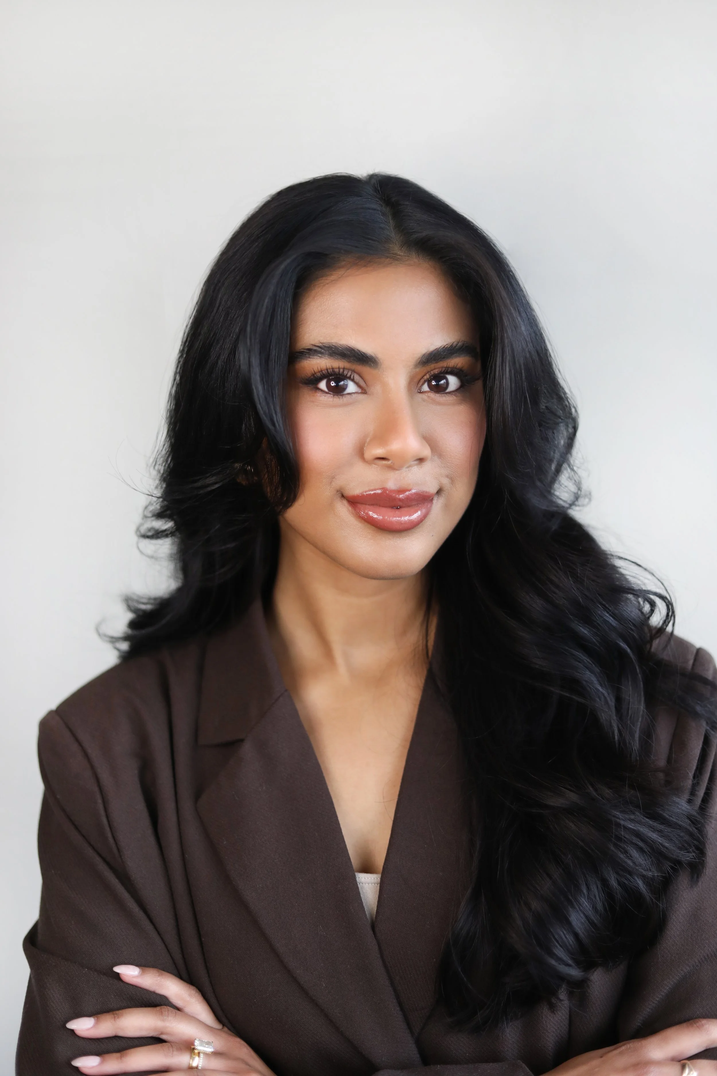 Close-up of a woman with long black hair, wearing a brown blazer, and posing with arms crossed, against a plain white background.