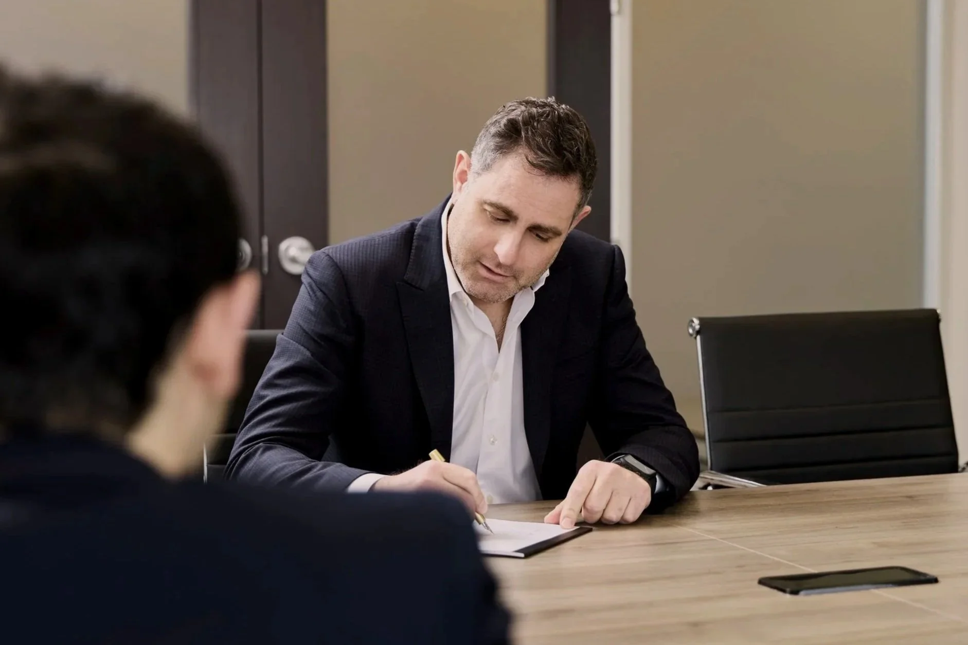 Man in a suit sitting at a conference table, writing notes, with a woman in the foreground.