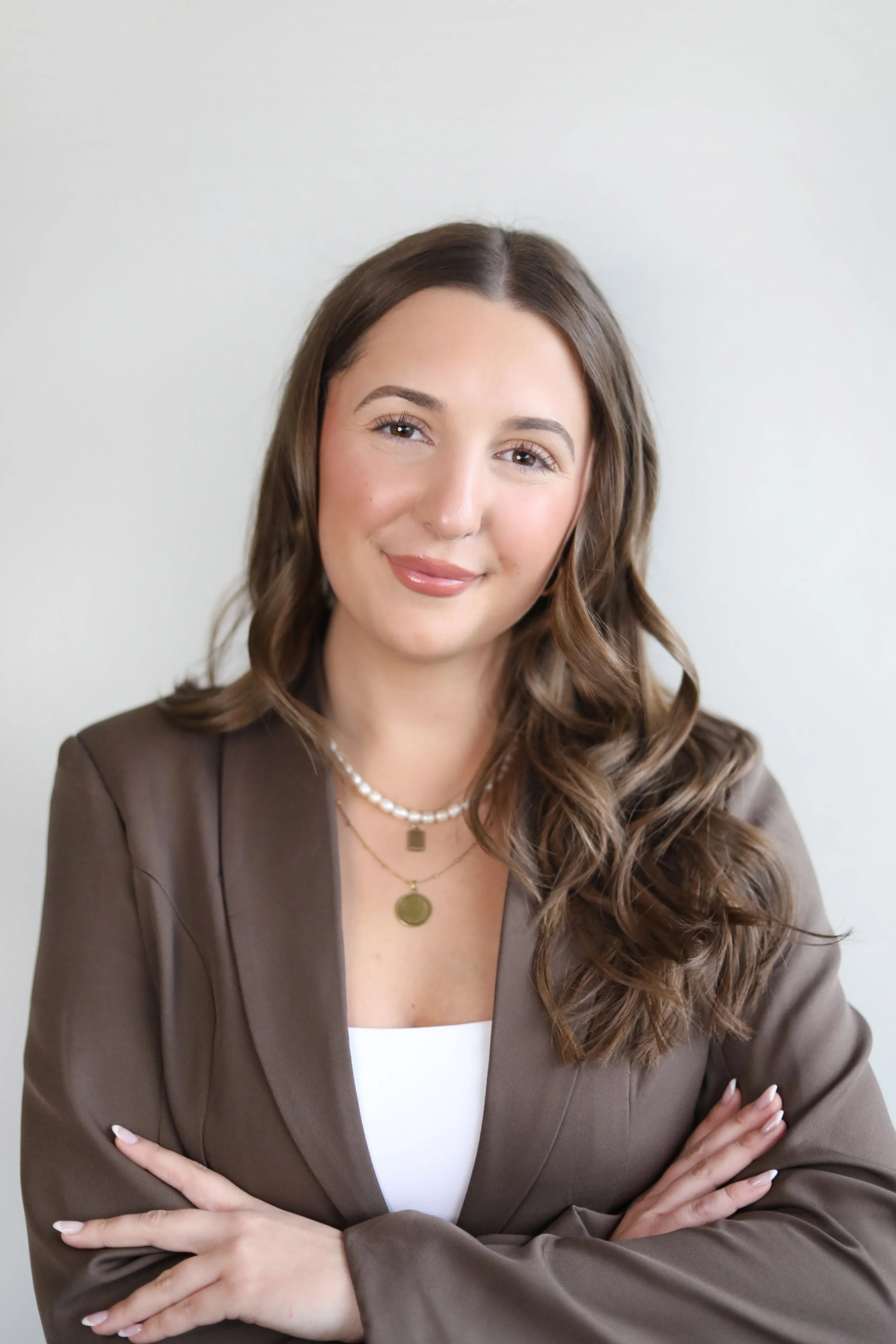 A woman with long, wavy brown hair, wearing a brown blazer, white top, pearl necklace, and layered gold necklaces, standing against a neutral background, with arms crossed and smiling gently.