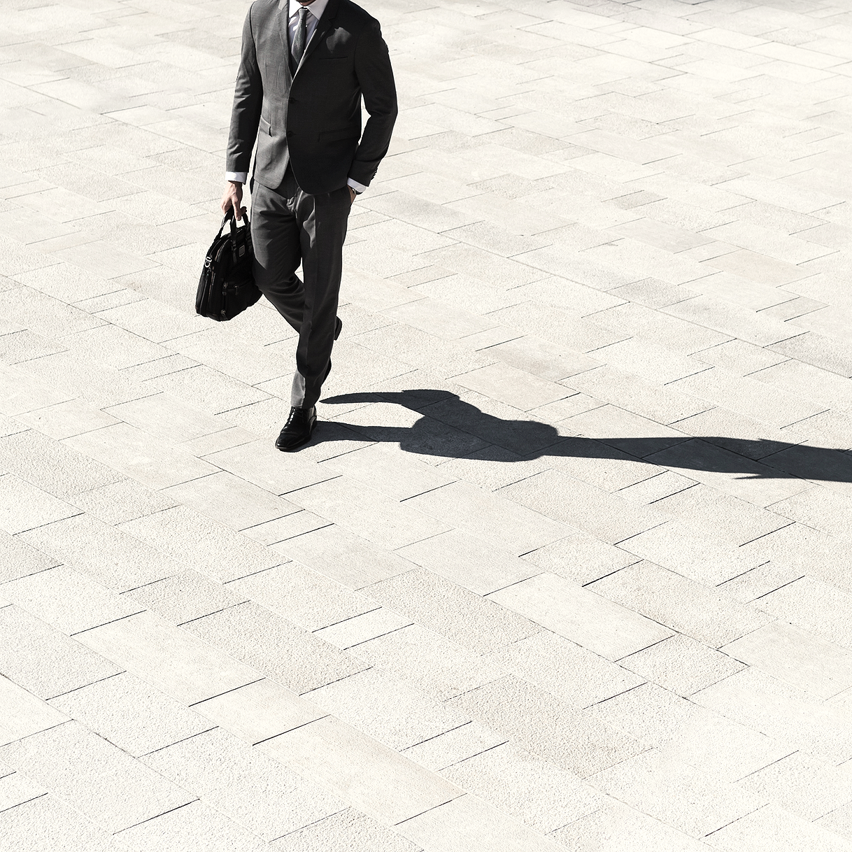 A man in a dark suit walking on a light-colored tiled surface, carrying a briefcase and casting a shadow.
