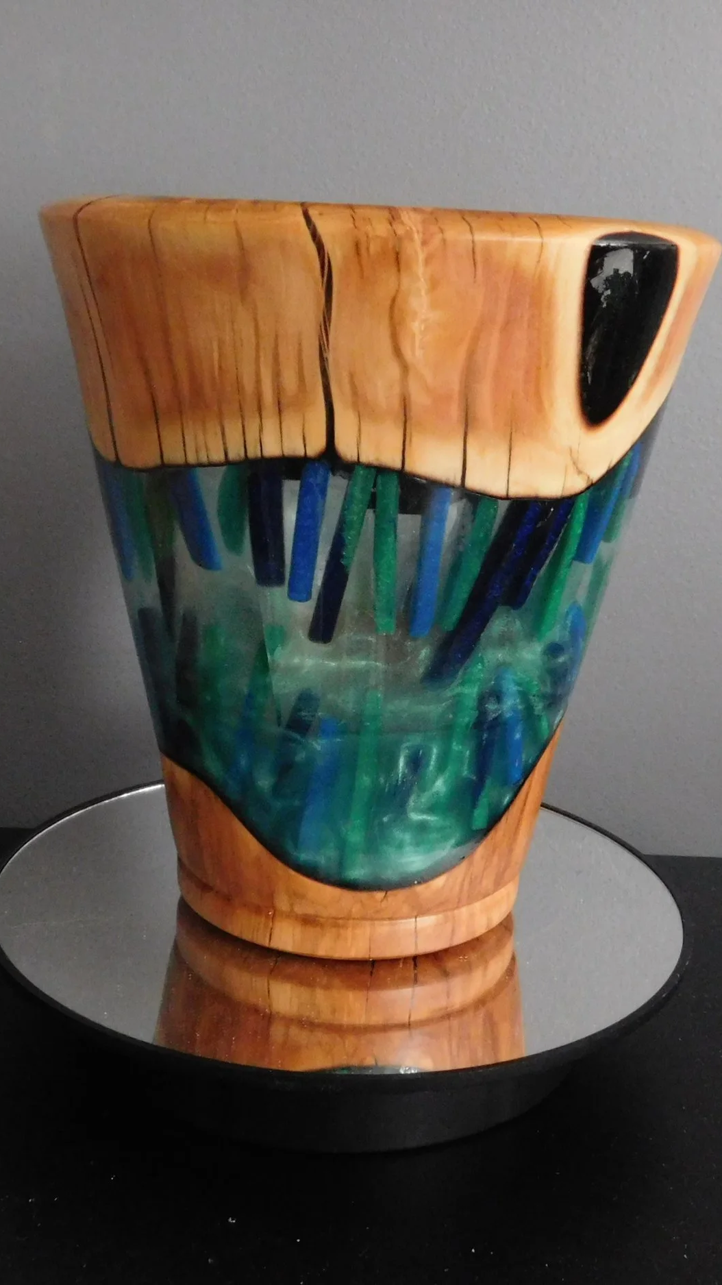 A unique ceramic mug with a wooden top, glass panel, and colorful glass handles, resting on a round mirrored tray.