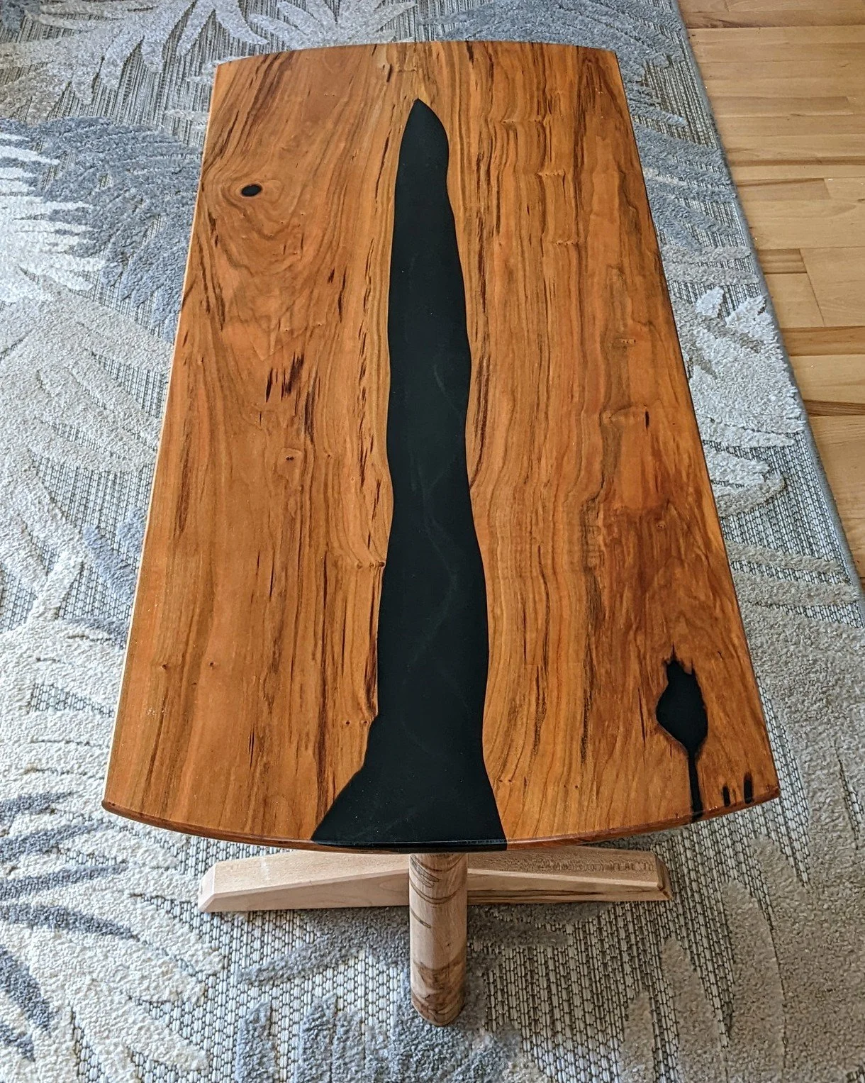 Wooden coffee table with black resin inlay in the center, placed on a rug with a wooden floor background.