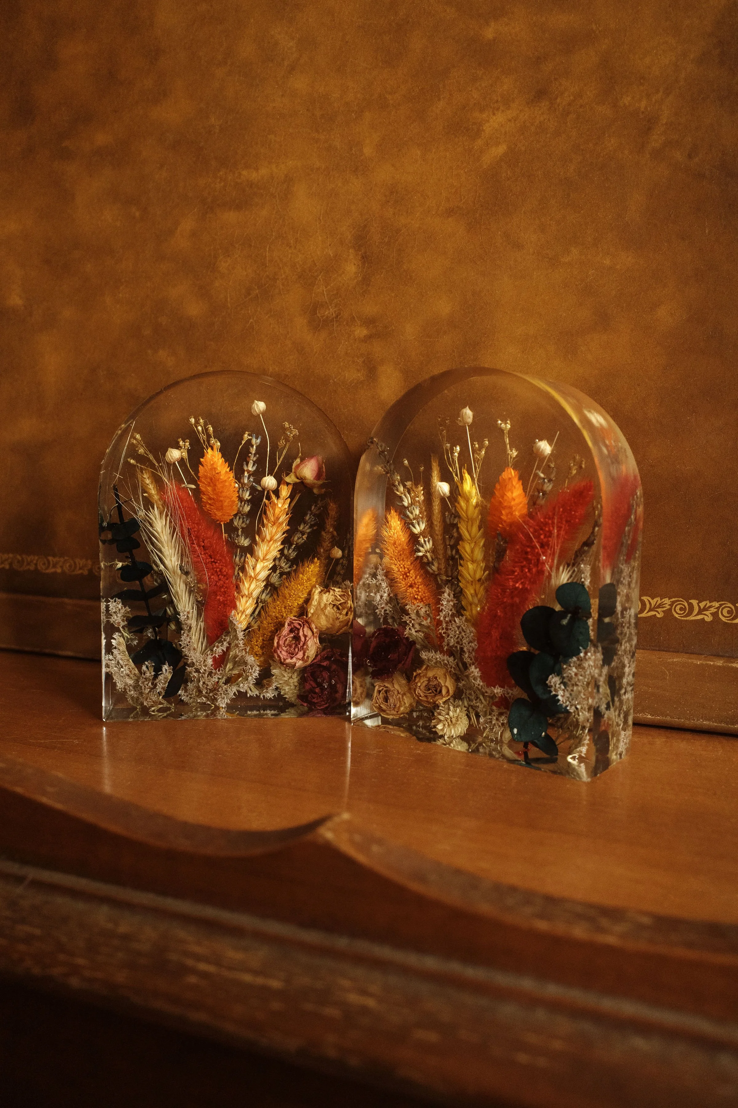 Two transparent acrylic blocks with preserved dried flowers and foliage, placed on a wooden surface in front of a brown textured background.