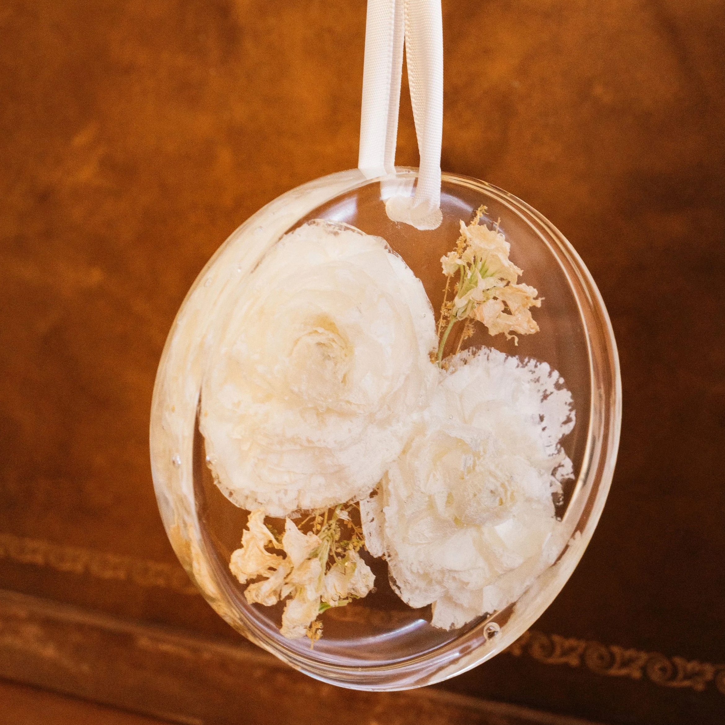 Round glass ornament with white roses and dried flowers inside, hanging by a white ribbon against a brown background.