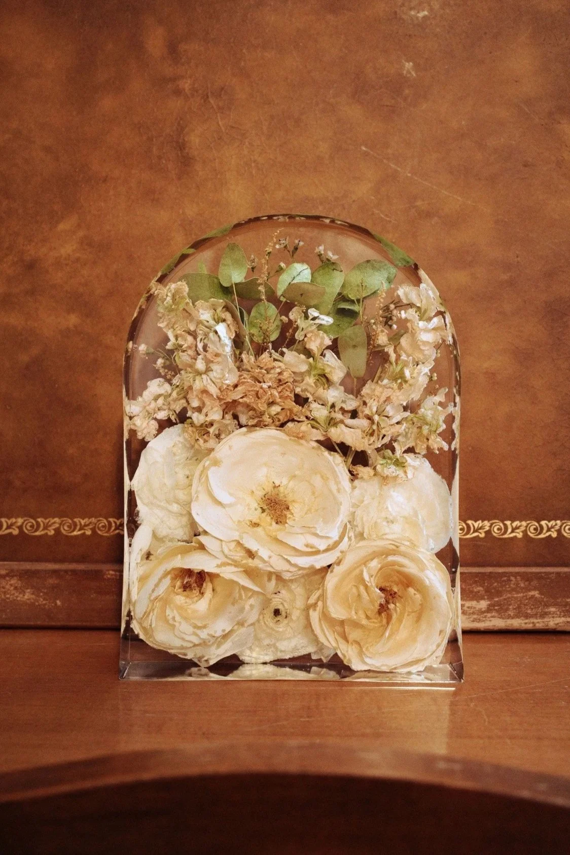 A clear glass display case with dried white flowers and green foliage inside, placed on a wooden surface against a textured brown background.