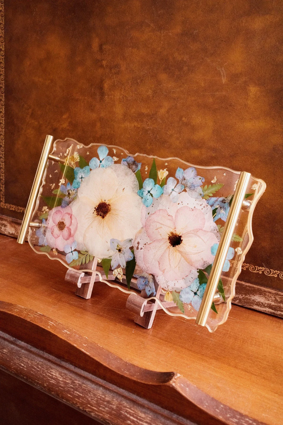 Decorative glass tray with pressed flowers, including pink, white, and blue blossoms, placed on a wooden surface against a brown background.