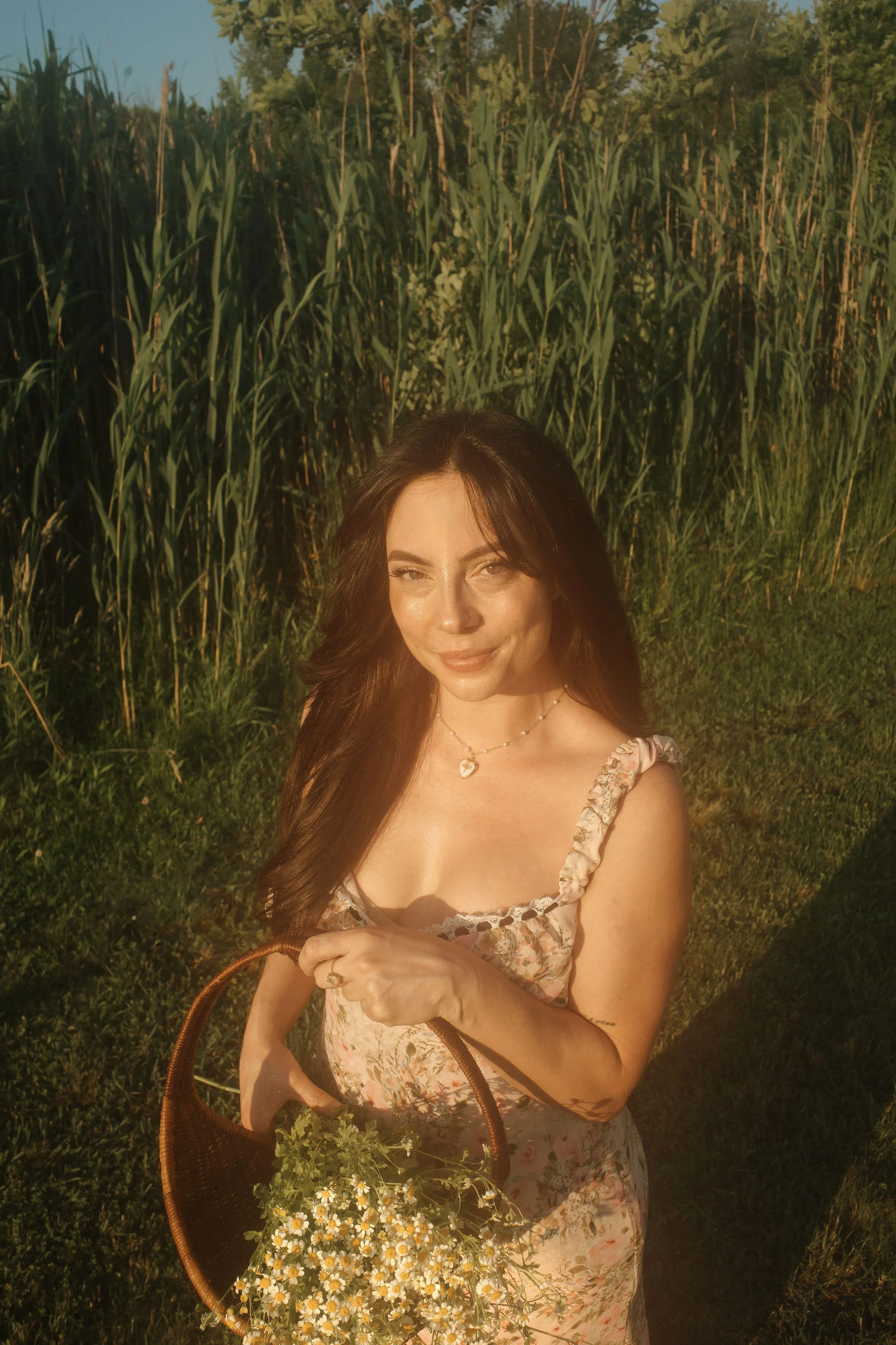 A woman holding a basket of daisies standing outdoors in a field with tall green crops, during sunset.