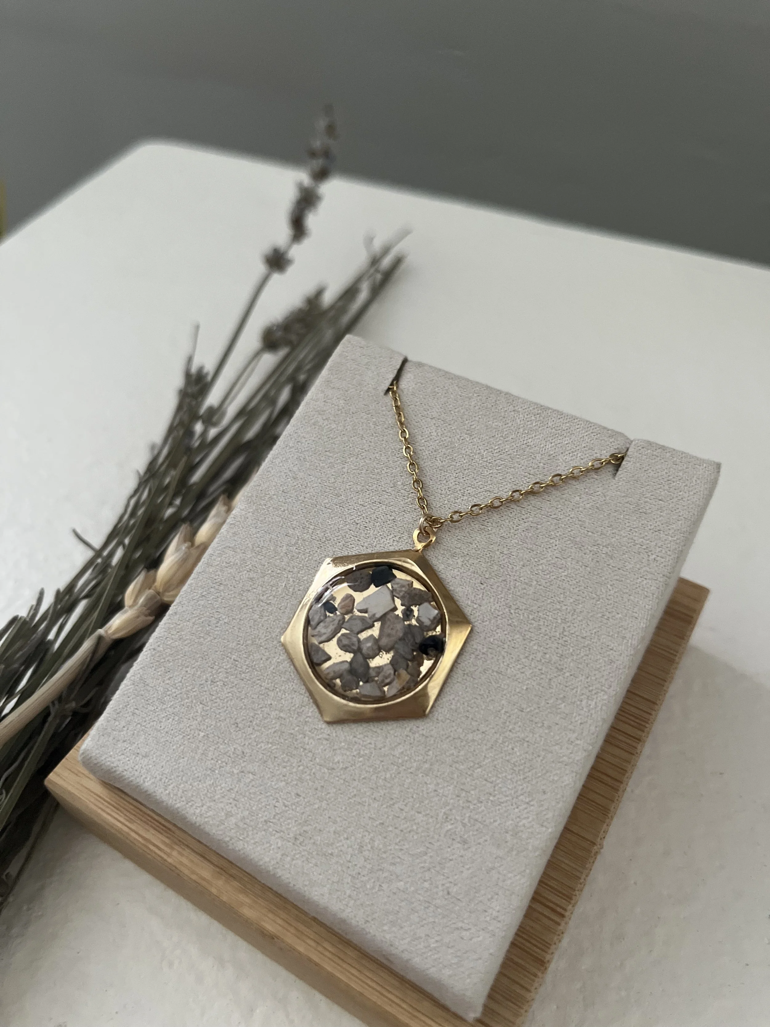 Gold necklace with a large, irregularly shaped pendant filled with small black, gray, and white stones displayed on a light grey jewelry stand.